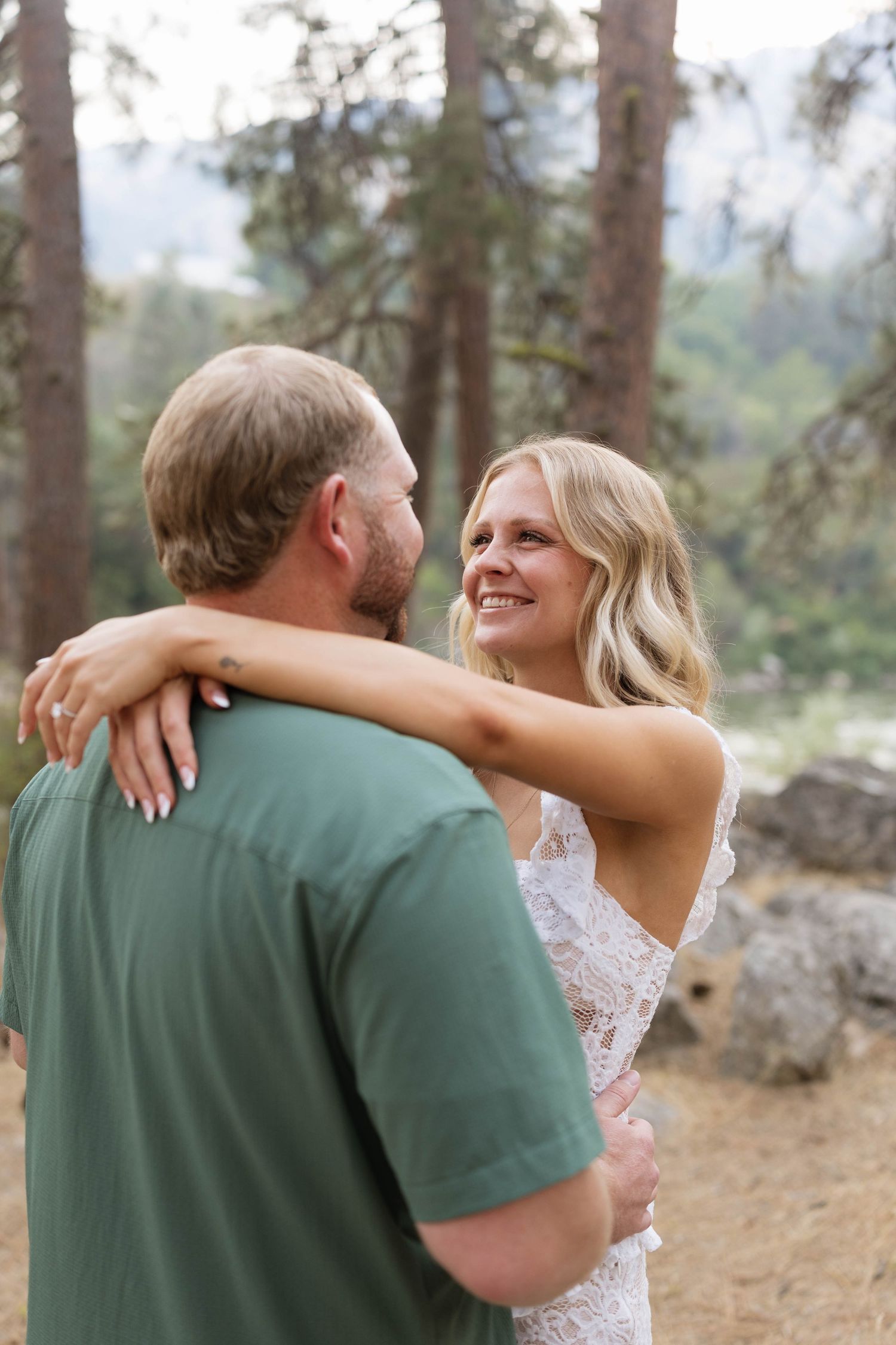 A smiling blonde woman in a lace dress embraces a man in a green shirt outdoors among pine trees.