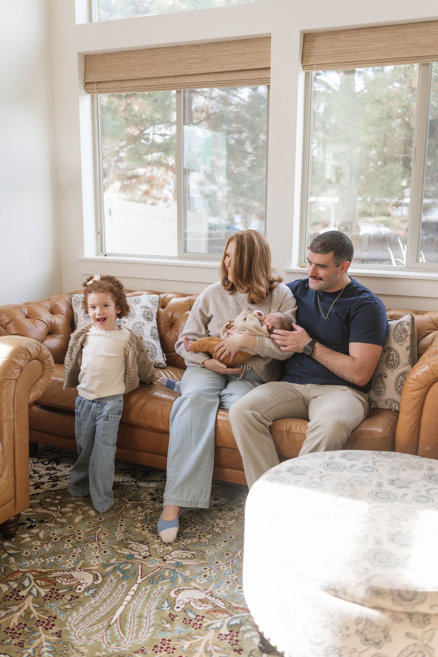 Family shares tender moments on a brown leather couch in a sunlit room with windows.