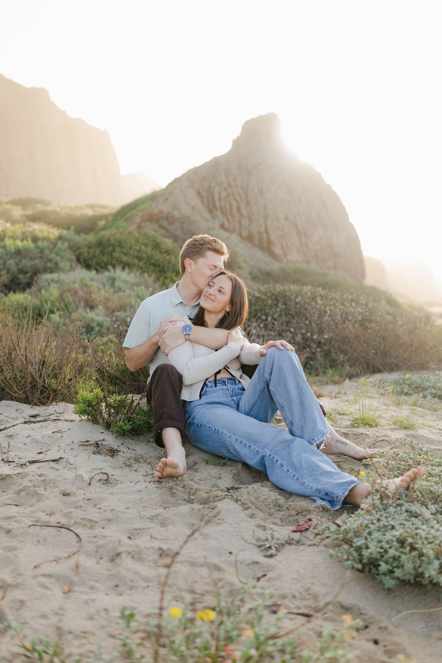 A couple shares a romantic moment together on a sandy beach at sunset with mountains in the background.