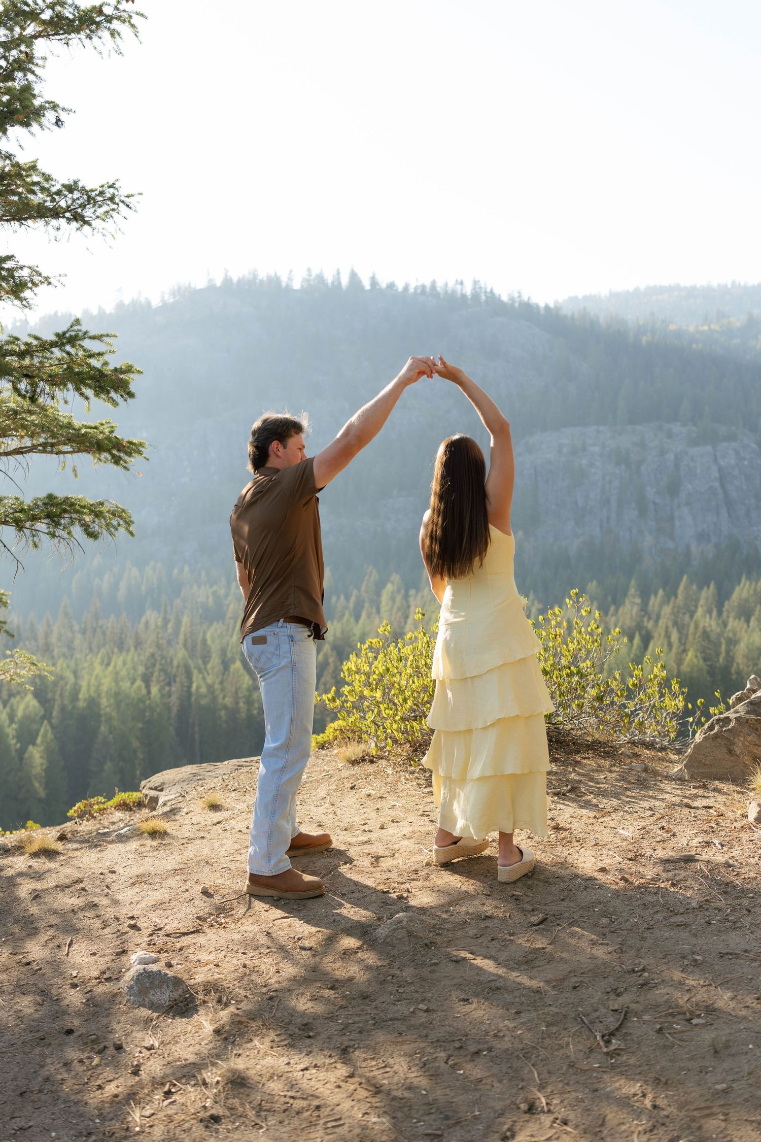 Two people stand together on a mountain overlook at sunset, raising their arms to form heart shapes against the forest vista.