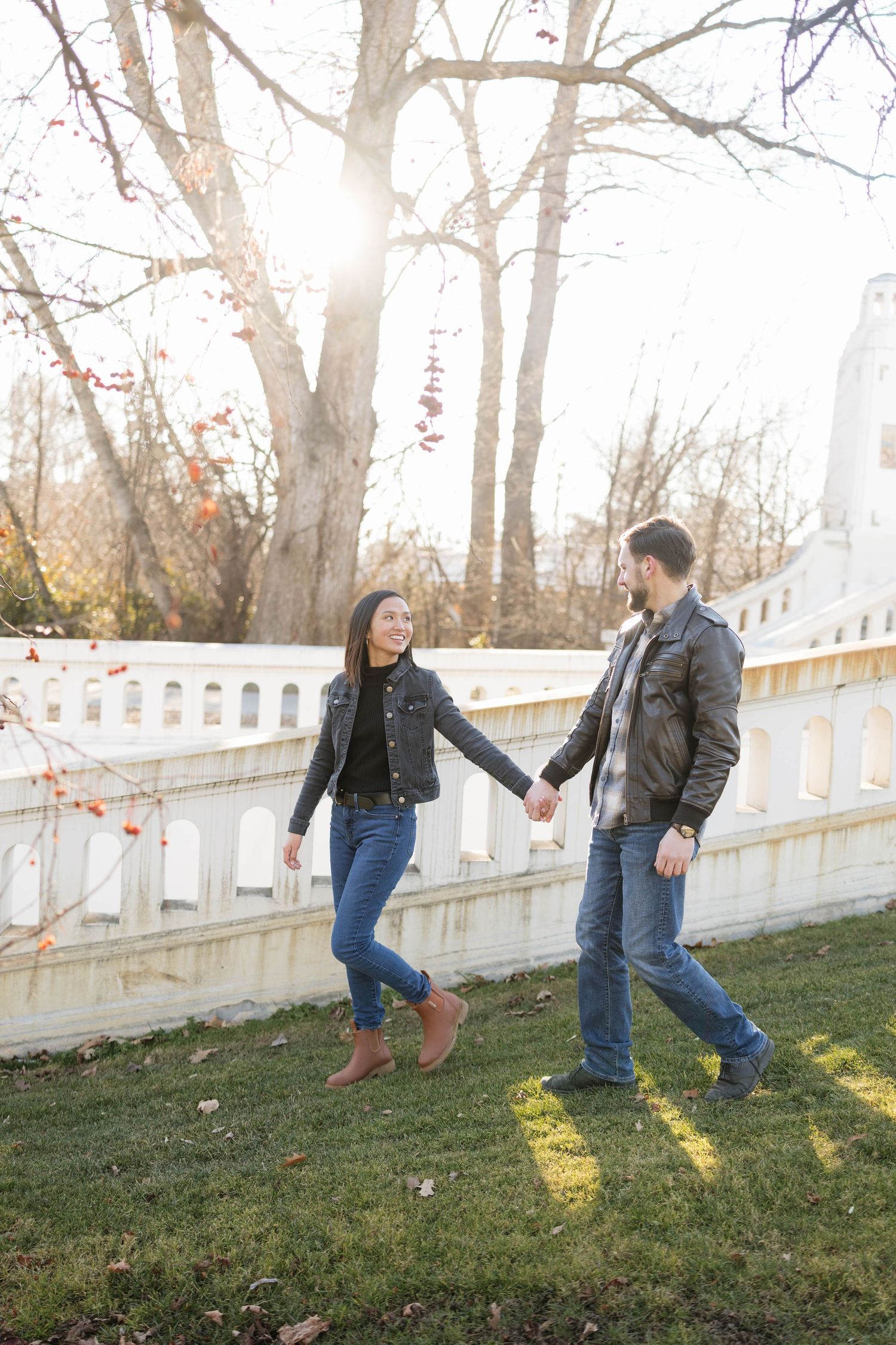 A couple holding hands and walking on grass near a white fence on an autumn day with sunlight streaming through trees.