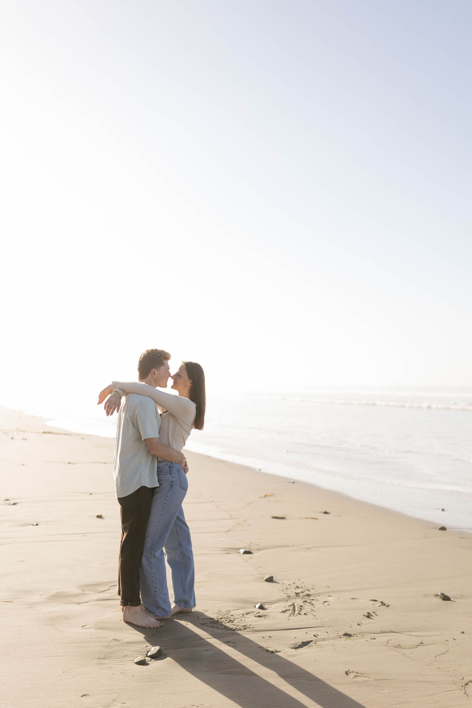 A romantic couple embraces on a sunny beach while casting long shadows on the sand during golden hour.