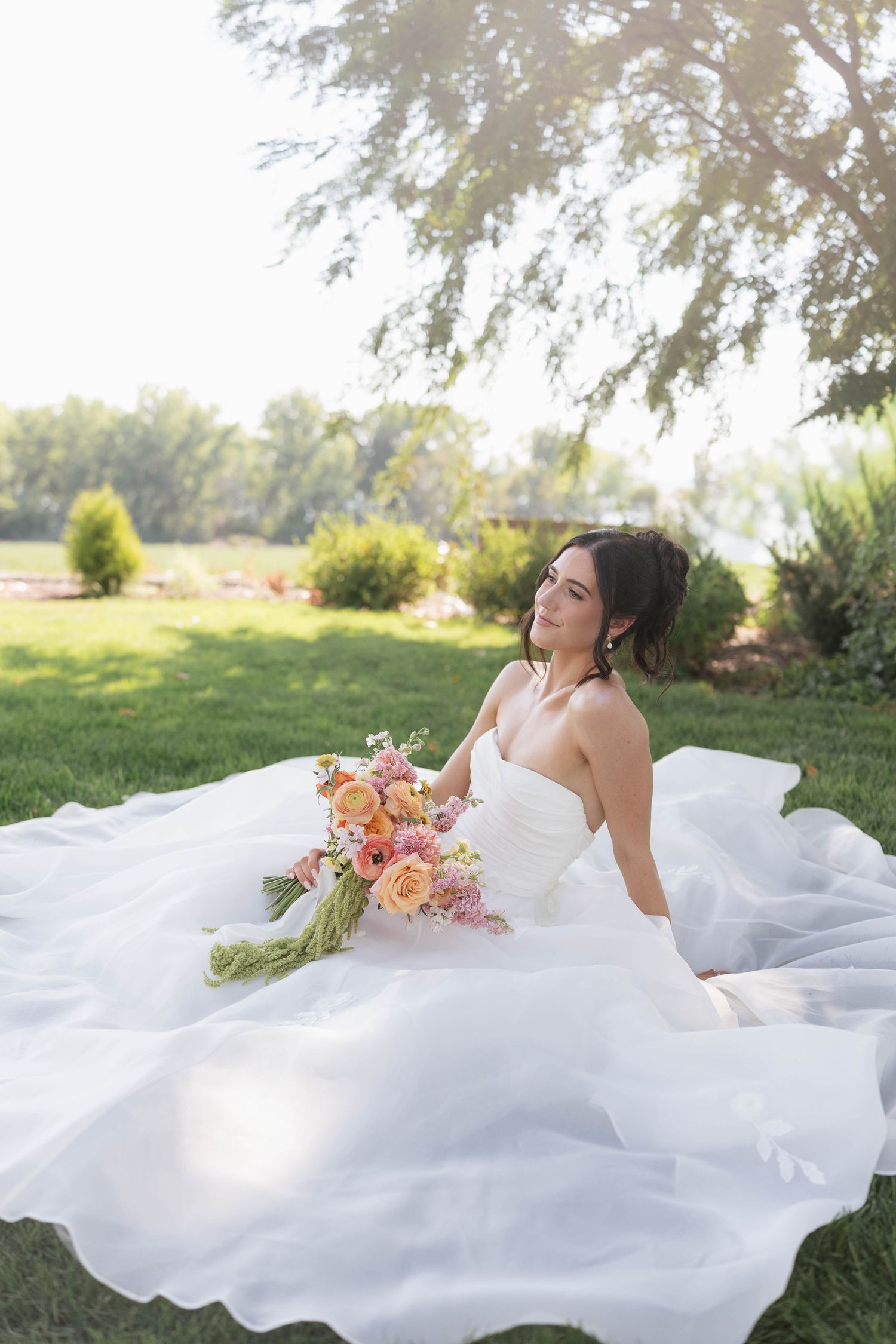 Bride in white gown sits on grass holding colorful pink and orange bouquet, with sunlit green trees in background.