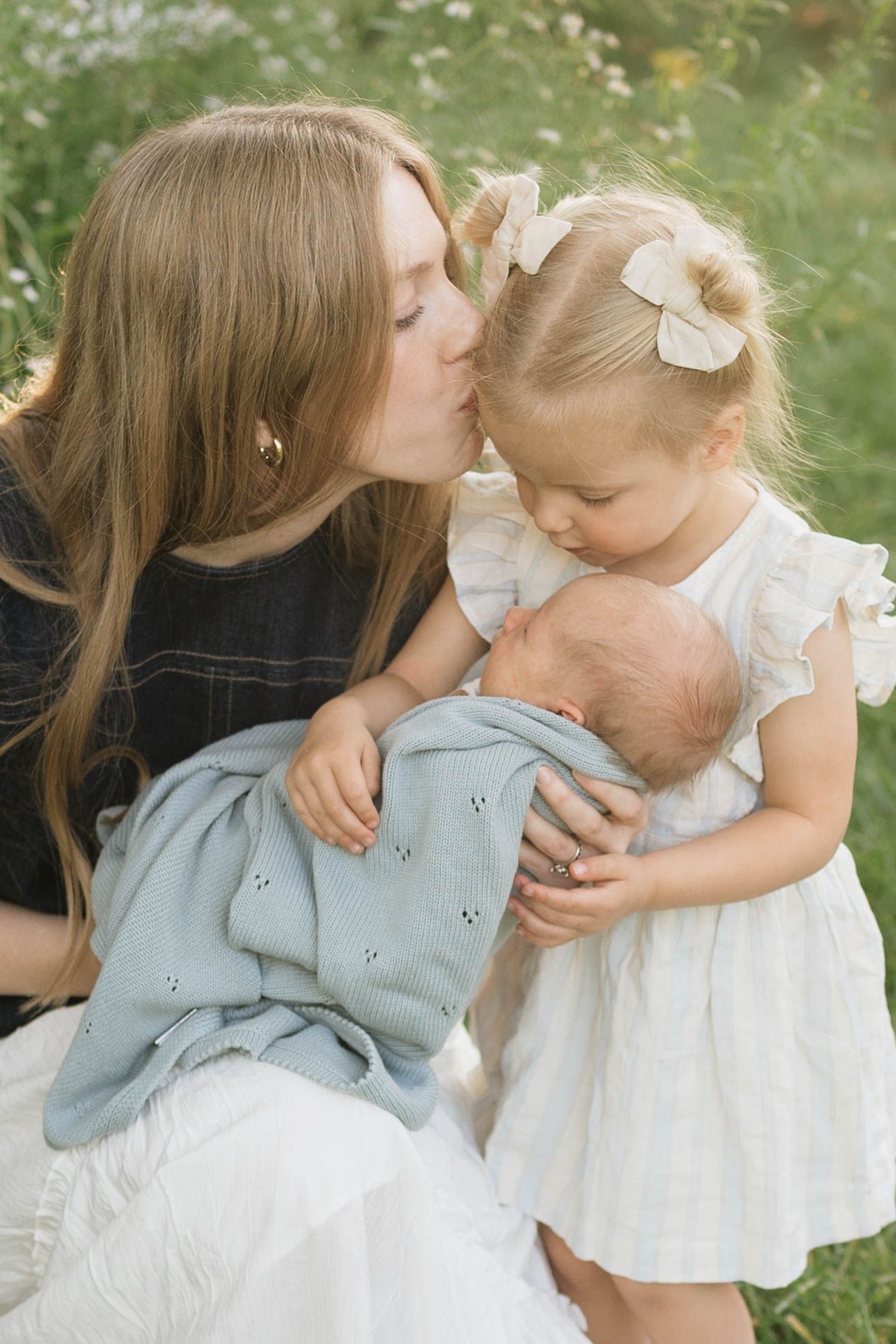 A mother kisses her toddler daughter while holding a newborn baby wrapped in a blue blanket outdoors.