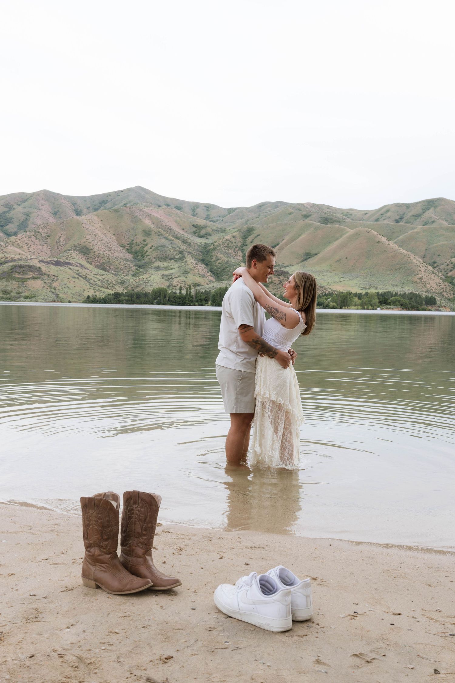Couple embracing in shallow lake water with cowboy boots and sneakers on sandy shore, mountains behind.