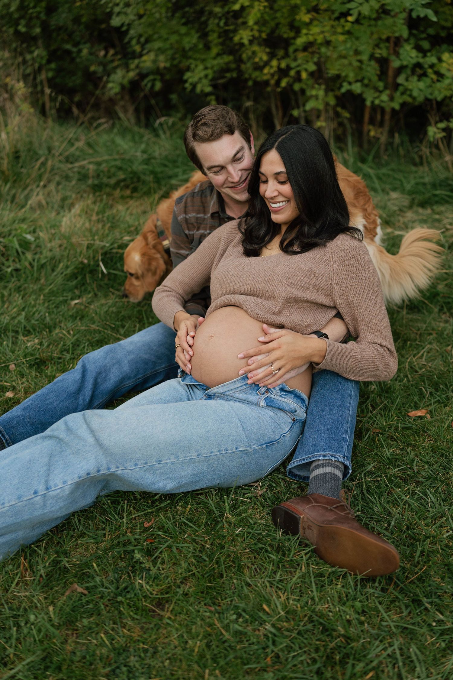 Smiling couple sitting on grass, pregnant woman cradling baby bump, man embracing her from behind, golden dog nearby.