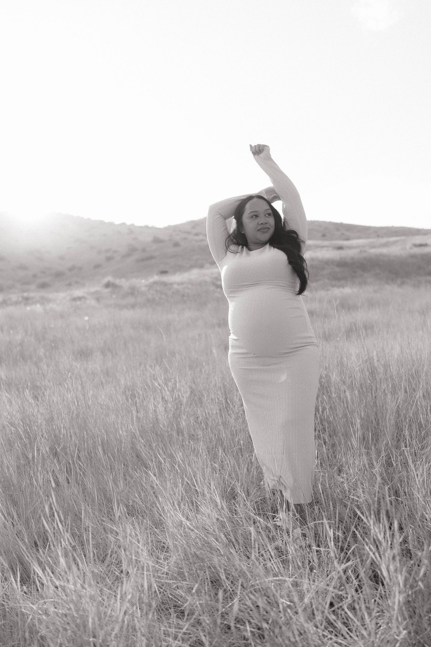 Black and white photo of a pregnant woman in a white dress posing in a grassy field with hills in the background.