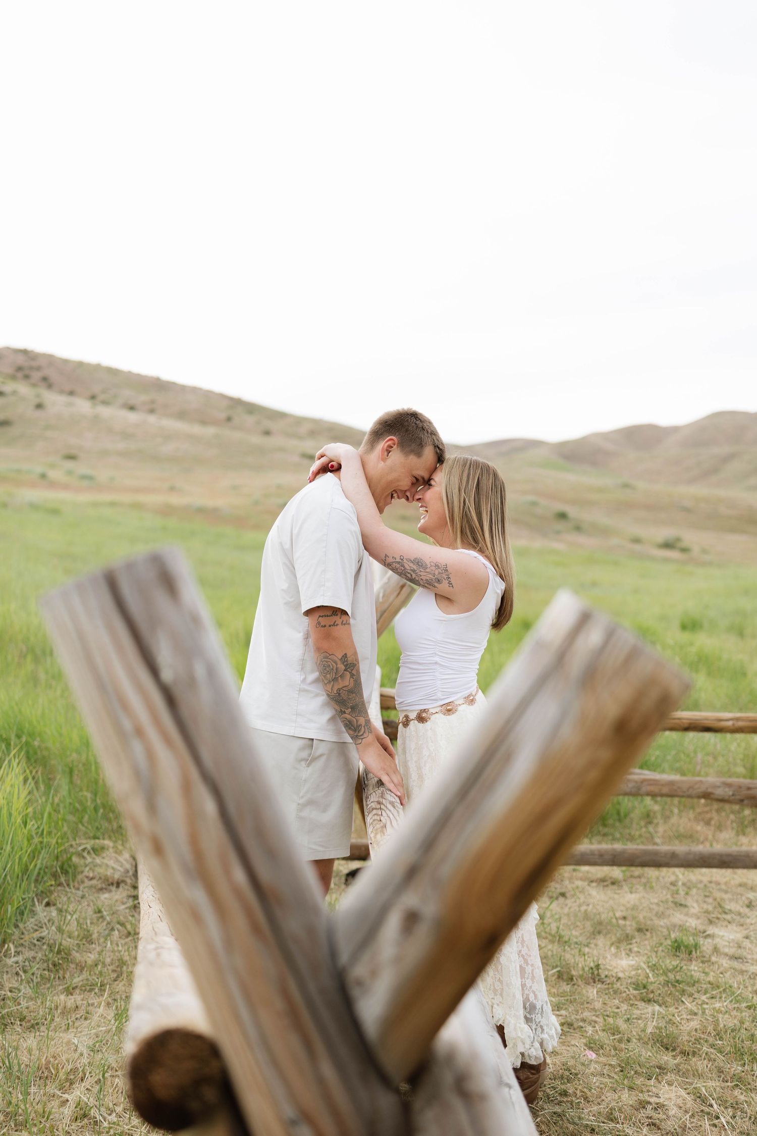 Couple with tattoos sharing a romantic moment near a rustic wooden fence in a green meadow with rolling hills.