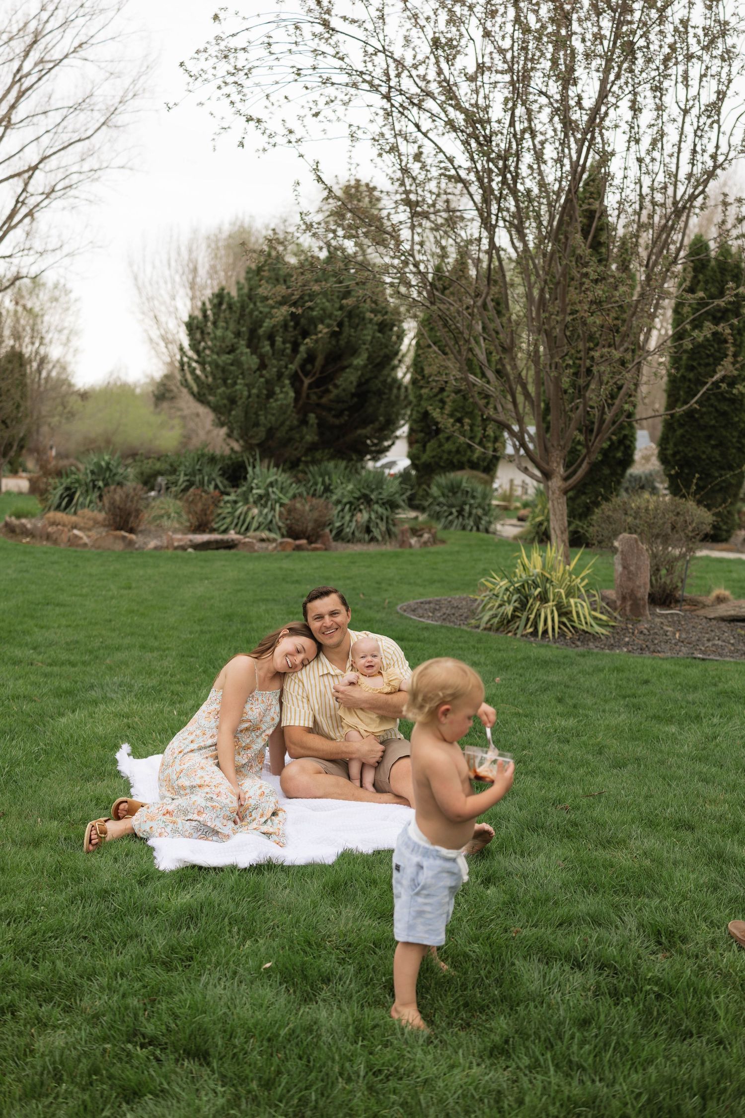 A family enjoys a relaxing afternoon picnic on a blanket in their backyard garden with trees and landscaping.