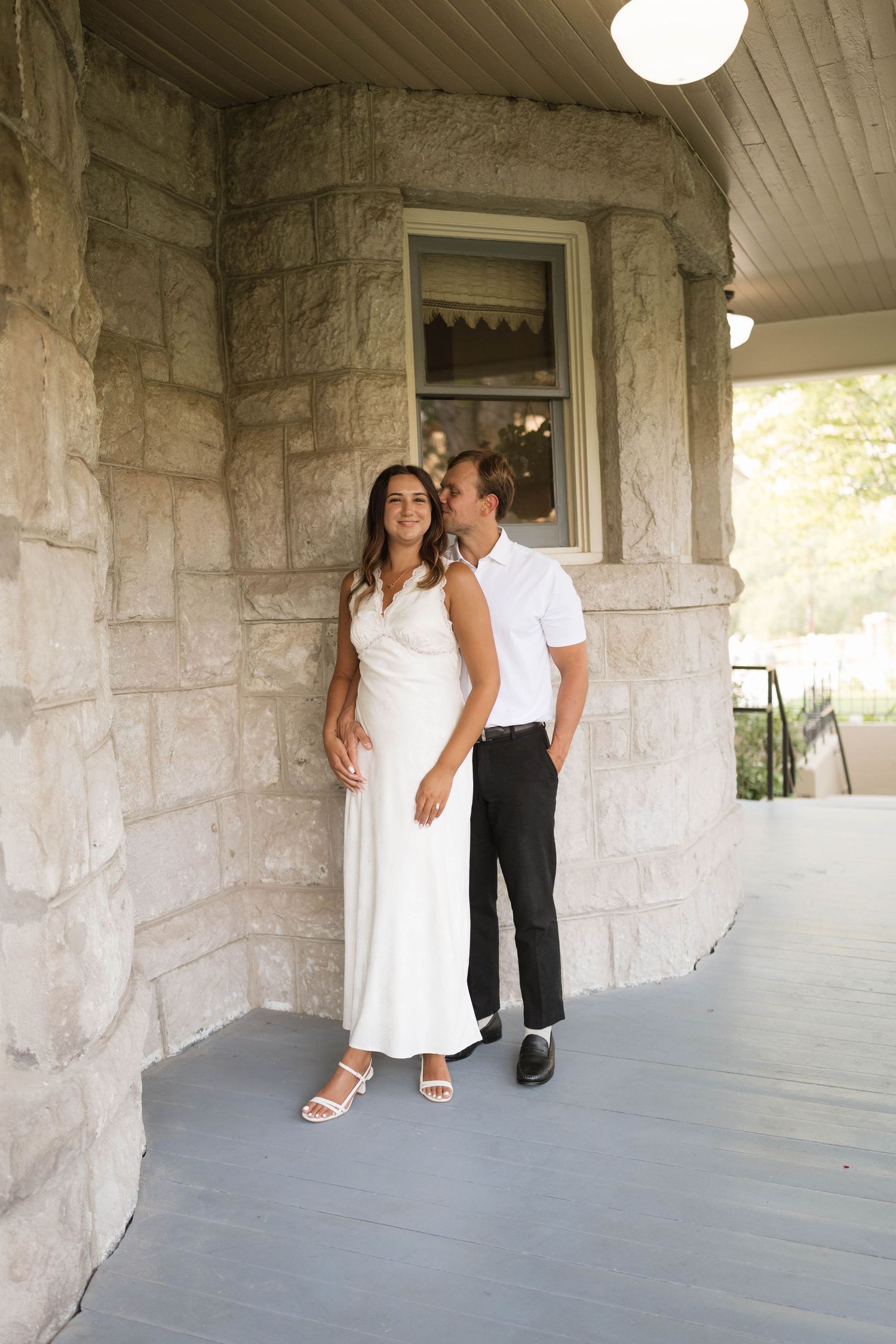 Couple in white dress and black pants posing on stone building porch for wedding engagement photos.
