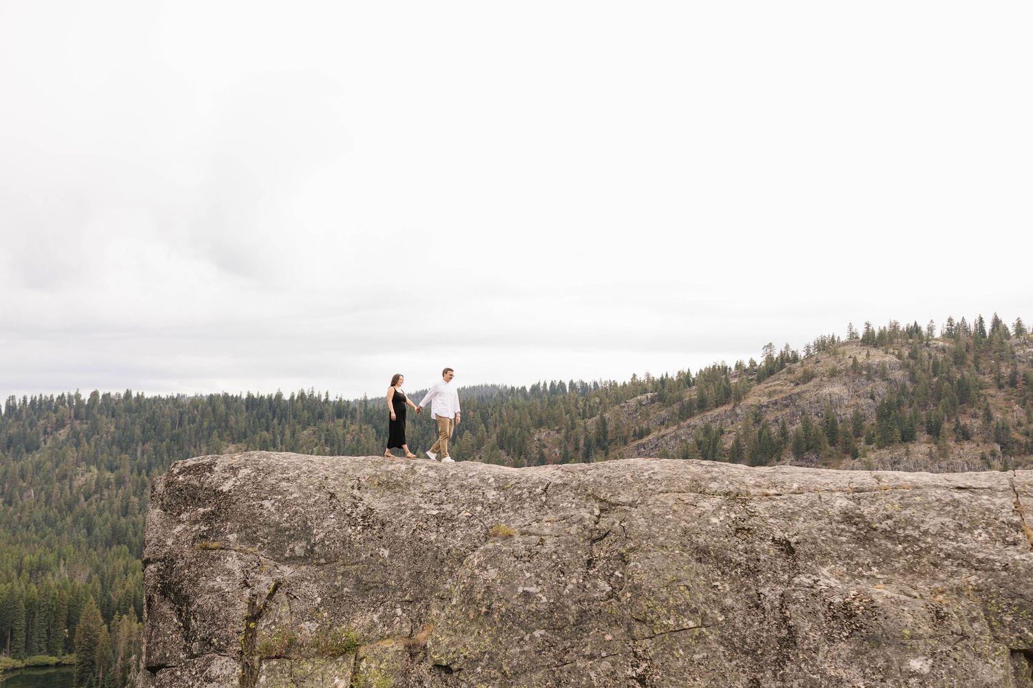 Couple standing on a rocky cliff overlooking a dense pine forest and mountains under an overcast sky.