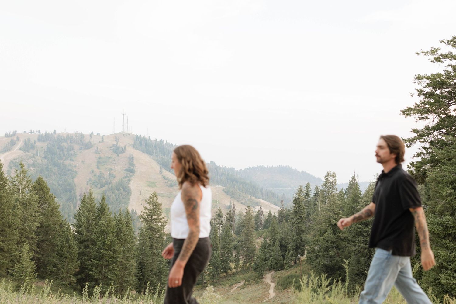 A couple walks along a grassy mountain trail surrounded by evergreen trees, with forested hills and a hazy sky in the background.