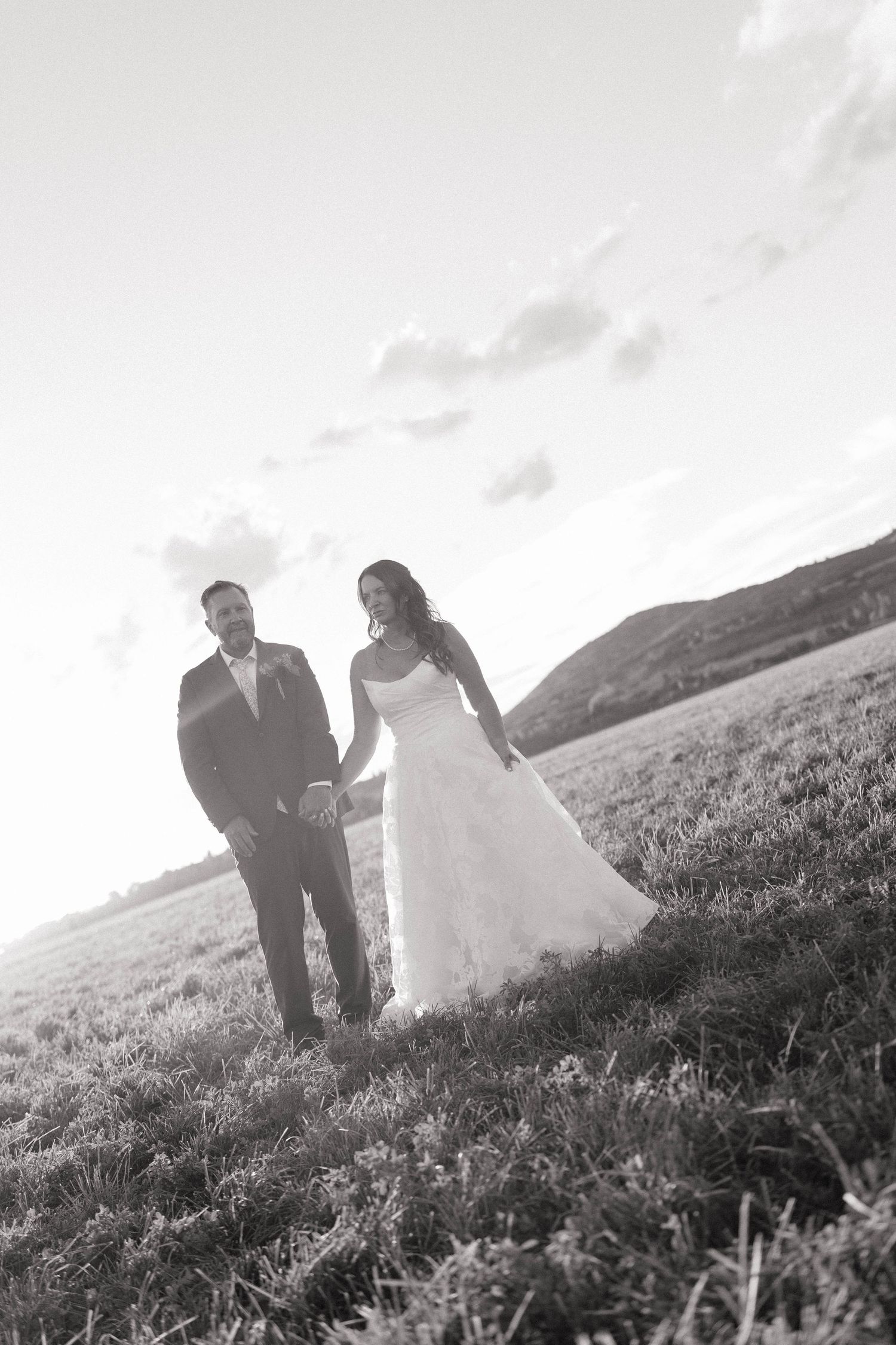 Black and white photo of a bride in a lace wedding gown holding hands with her groom in a suit on a grassy hillside.