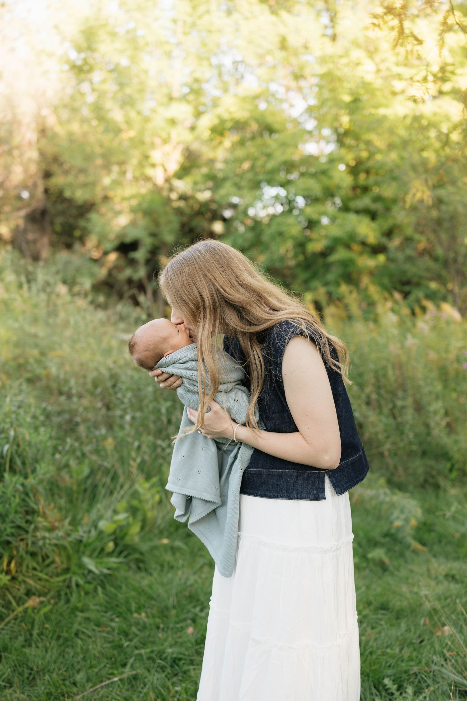 Mother in a white skirt and denim vest kisses her newborn baby wrapped in a sage blanket while standing in a sunlit green field.