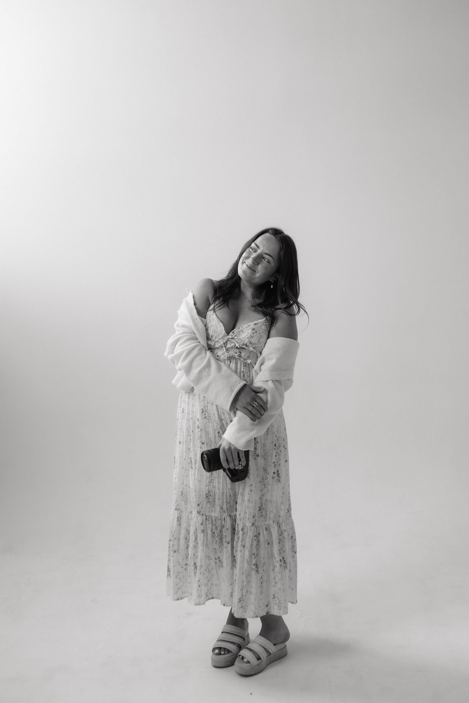 Black and white portrait of a woman in a floral dress and white cardigan holding a camera, posing in a studio.