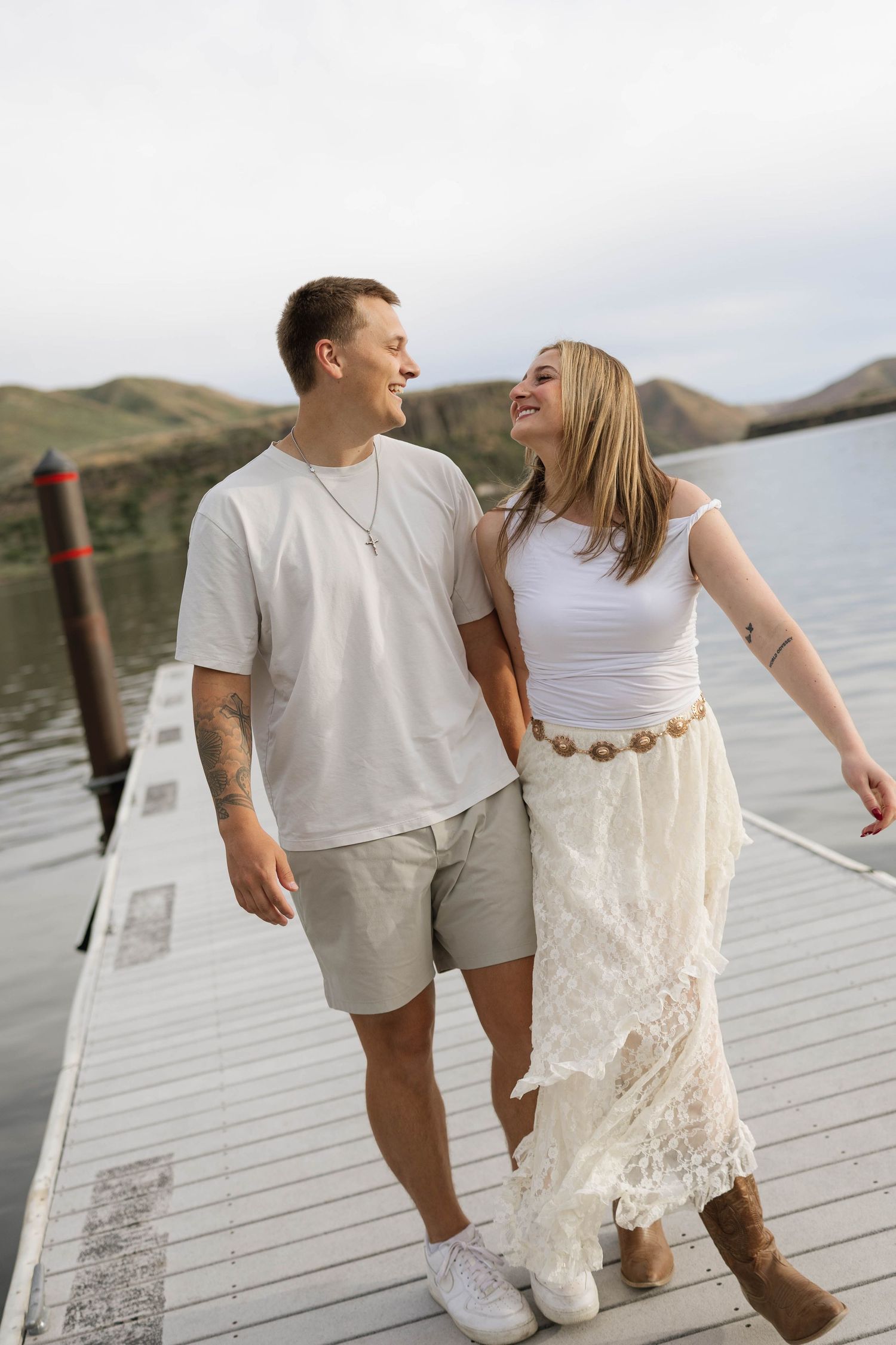 A smiling couple walks hand-in-hand on a dock by a lake, man in white tee and shorts, woman in white top and lace skirt.