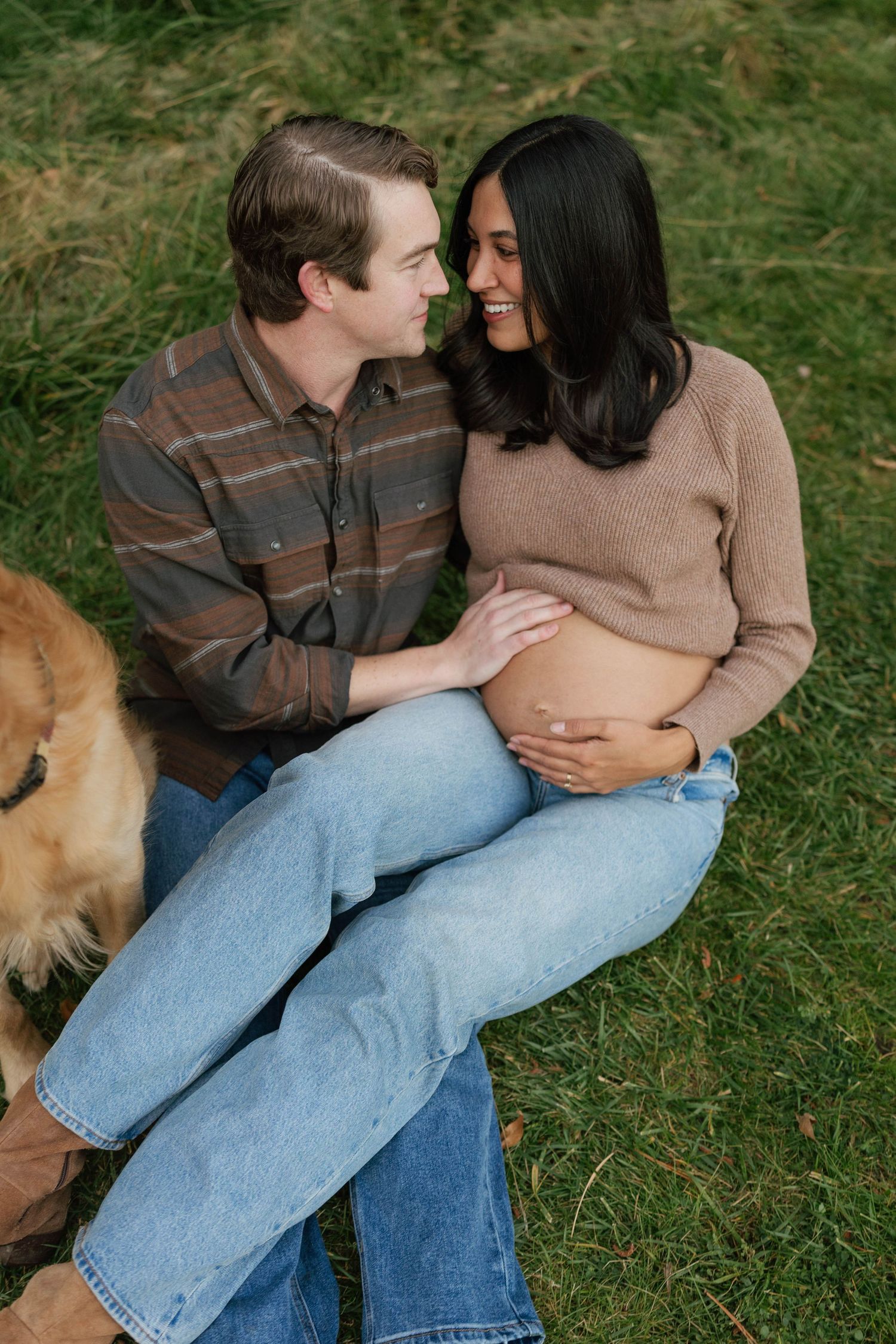 Couple sits together on grass sharing intimate moments while enjoying nature outdoors. Maternity session with golden retriever.
