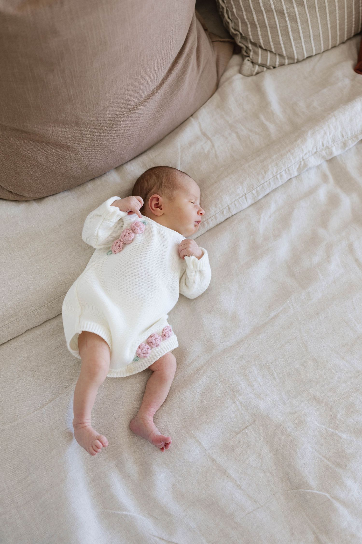Newborn baby sleeping peacefully on a linen bedspread in a white knit romper with pink floral details, beside neutral pillows.