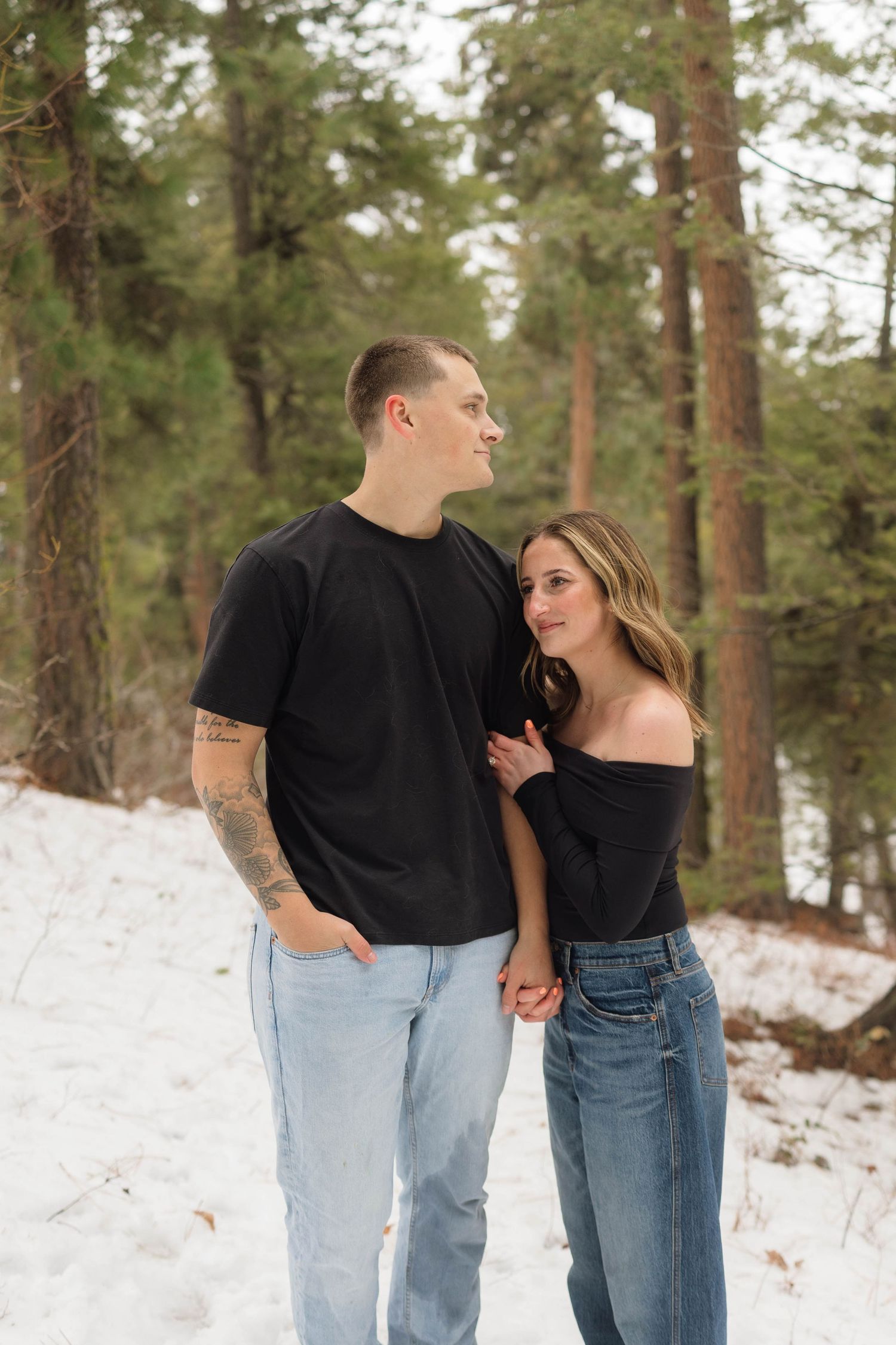 Romantic winter forest portrait of couple in casual outfits standing together in snowy pine woods.