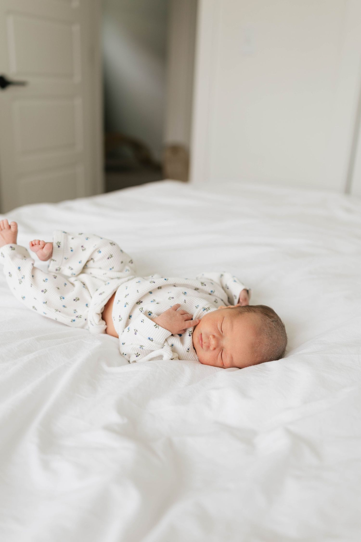 A newborn baby sleeps peacefully on white bedding wearing a patterned onesie.