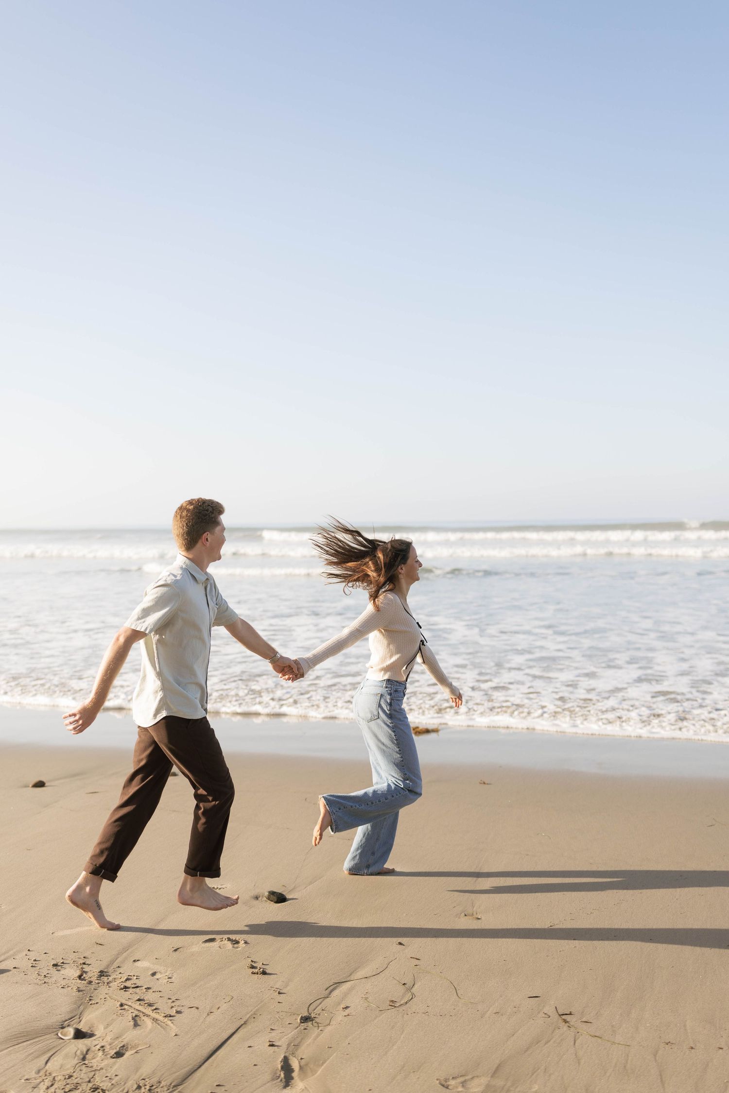 Two people playfully run and hold hands together on a sunny beach with waves crashing in the background.