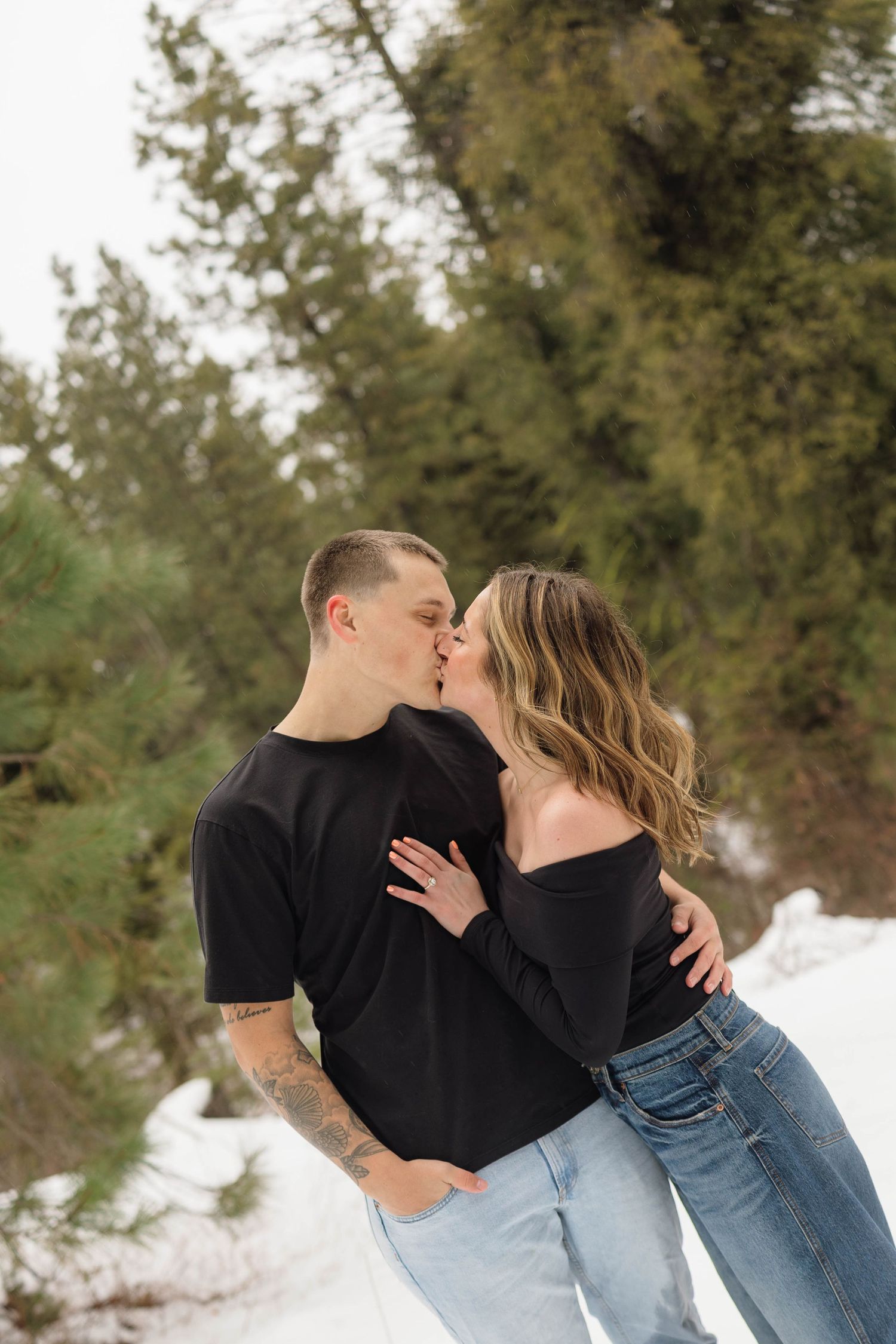 Romantic couple embraces in a snowy forest setting wearing casual winter outfits.