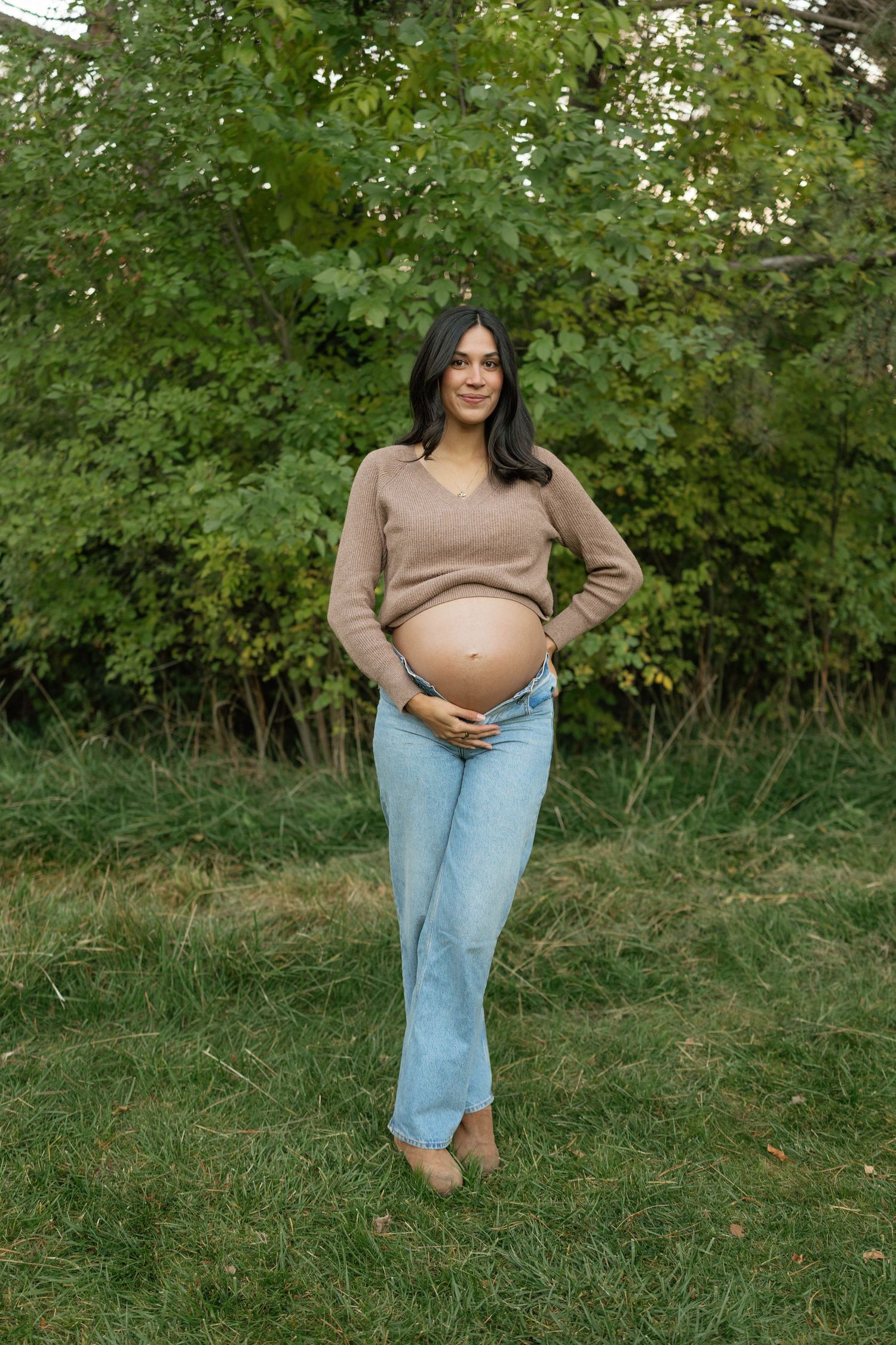 A maternity photoshoot features casual styling with jeans and a brown top against a natural green backdrop.