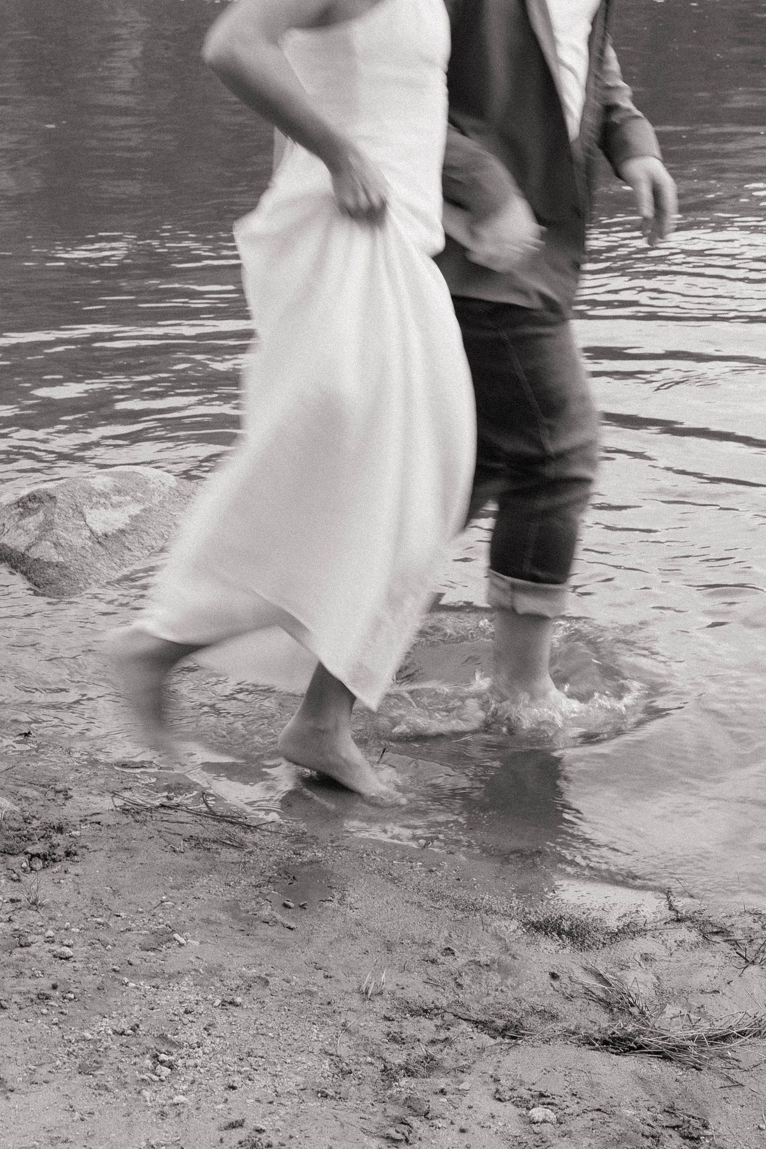 Black and white photo of a bride and groom walking barefoot through shallow water, wedding dress lifted.