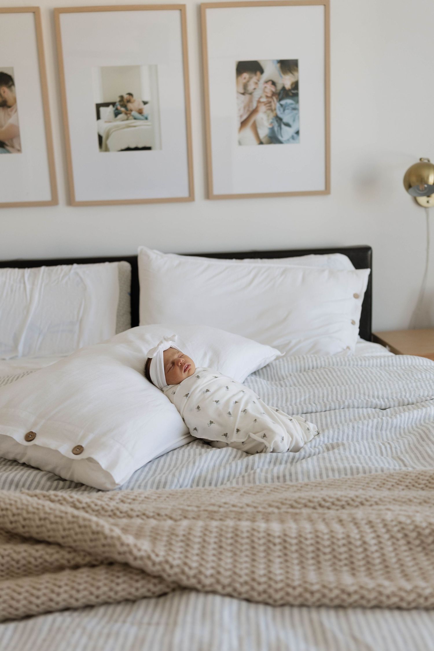 Newborn baby swaddled in white on a striped bed with framed family photos on the wall behind.