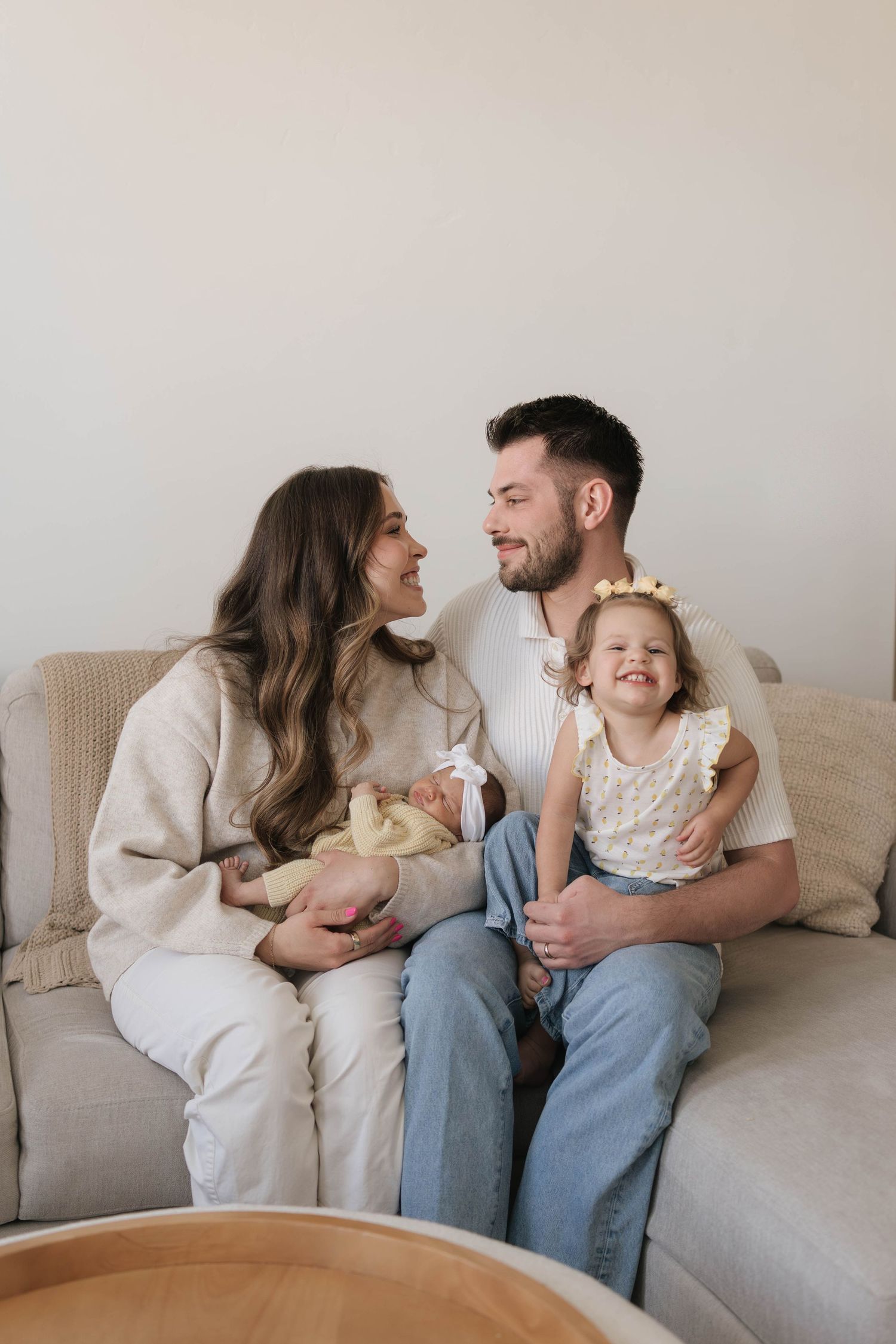 A happy young family sits on a beige sofa, parents smiling at each other while holding a newborn and a toddler girl.