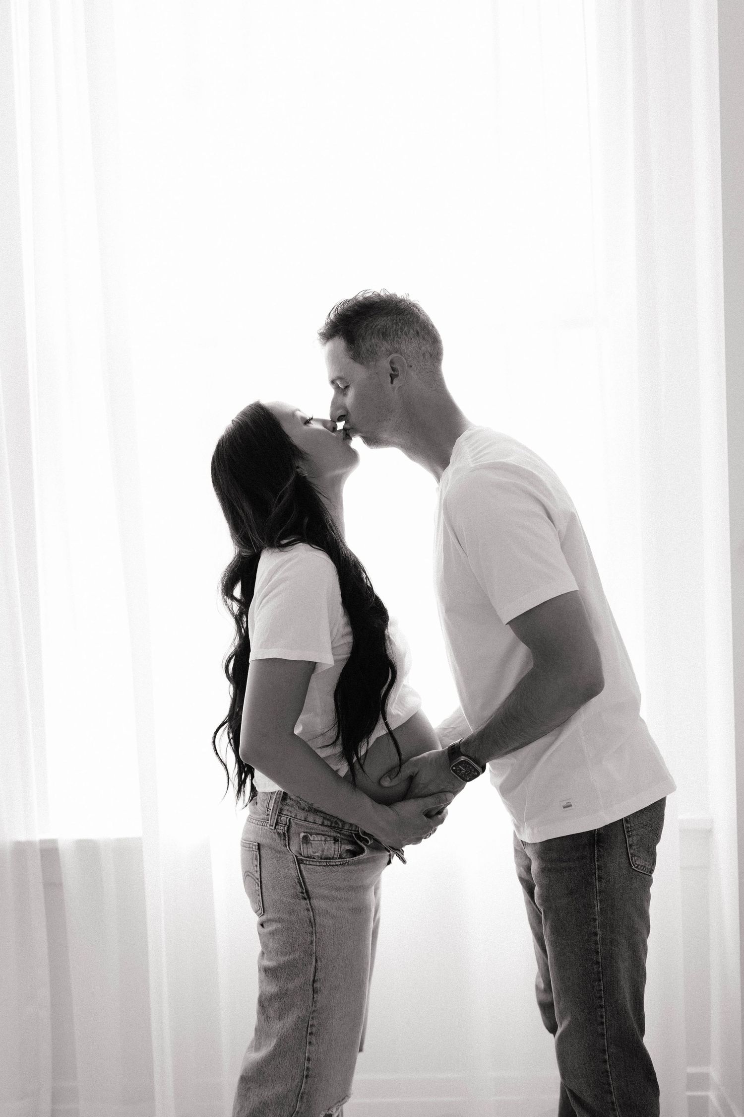Black and white photo of a couple sharing a tender kiss near sheer curtains, woman with long dark hair.