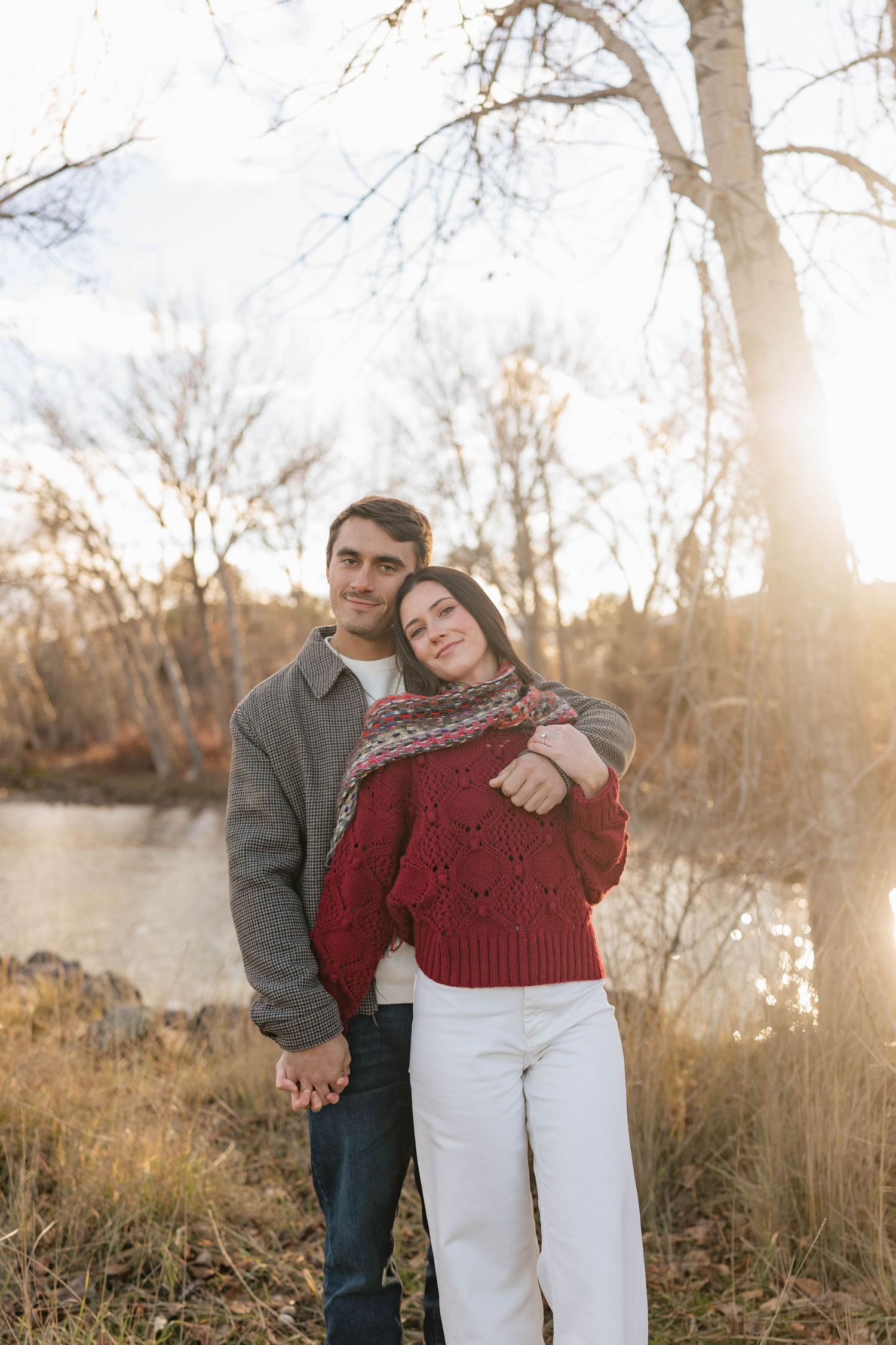 A romantic couple poses together in a natural setting with golden sunlight filtering through bare trees.