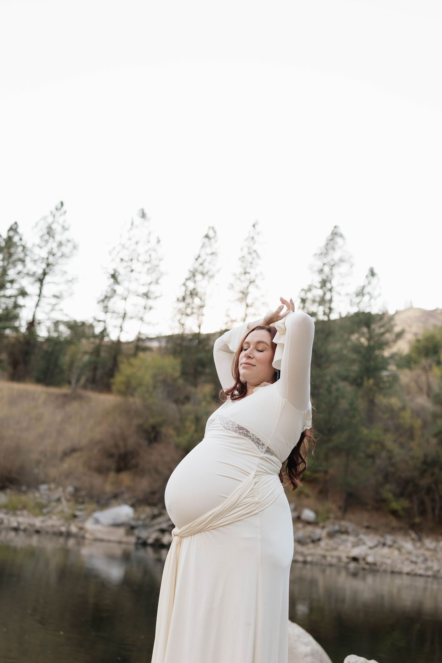 Pregnant woman in a flowing cream maternity gown poses with arms raised beside a calm river, with pine trees and hills behind her.