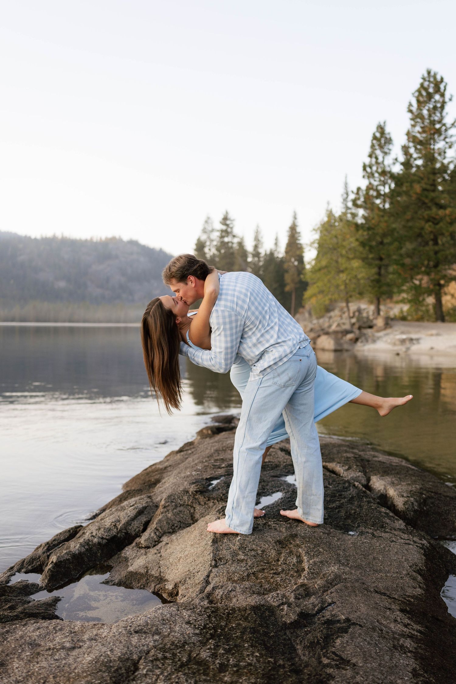 A couple shares romantic moments on large rocks by a serene lake surrounded by pine trees in the mountains.