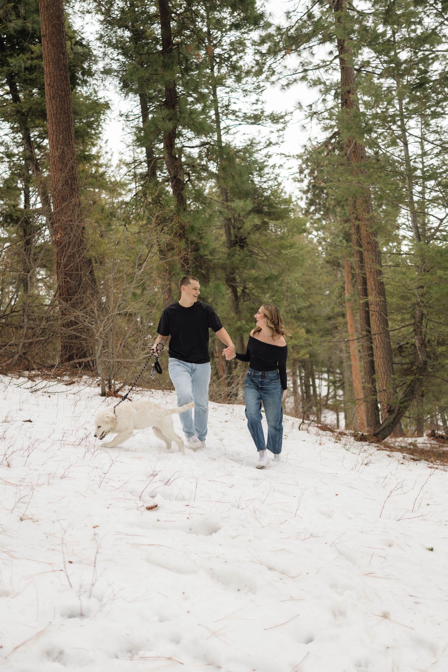 Two people walk together through a snowy forest path surrounded by tall pine trees on a winter day.