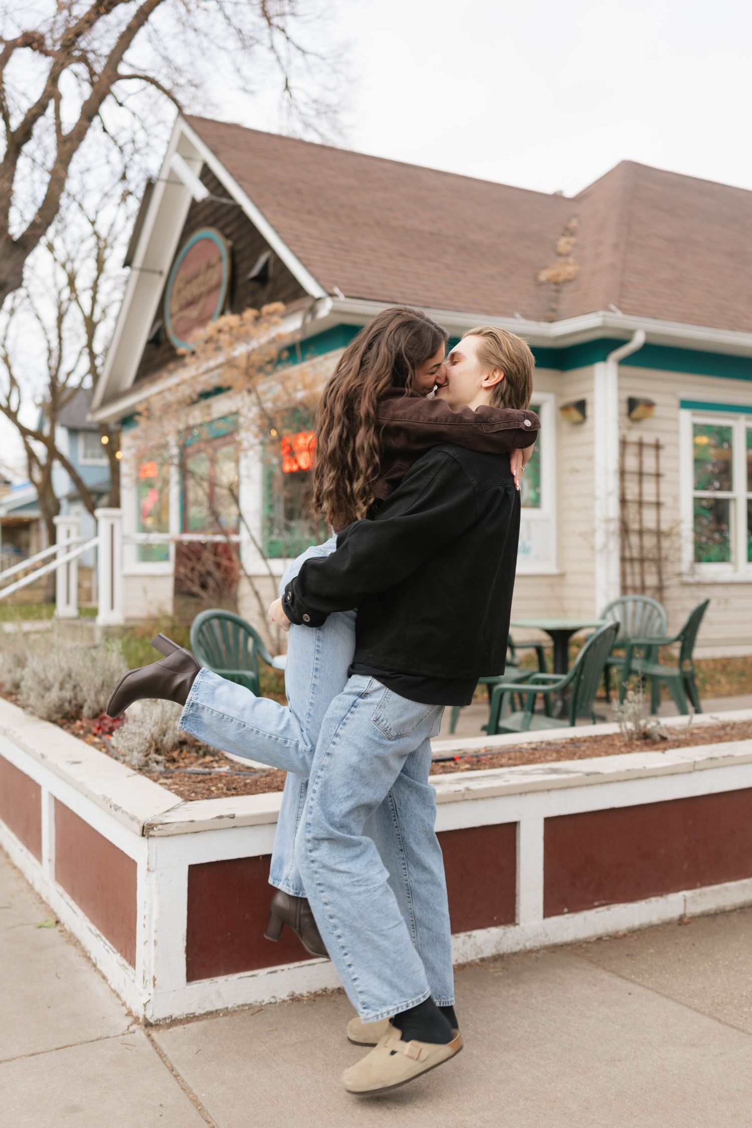 A couple embraces in a romantic moment outside a charming cottage-style house with a brown roof and green trim.