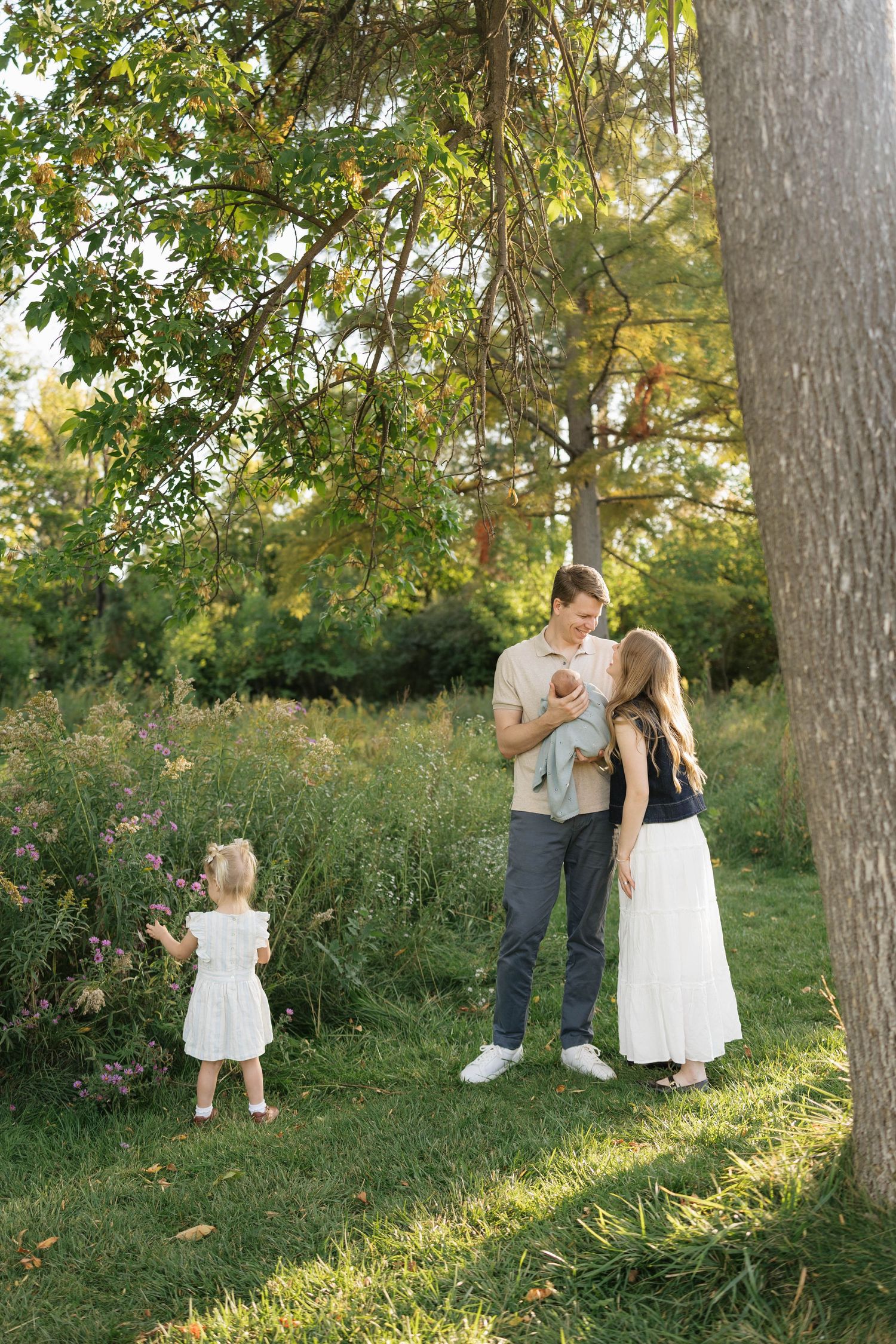 A family of four enjoys a sunny day outdoors, with parents holding a newborn while a toddler girl stands nearby in a lush green park.