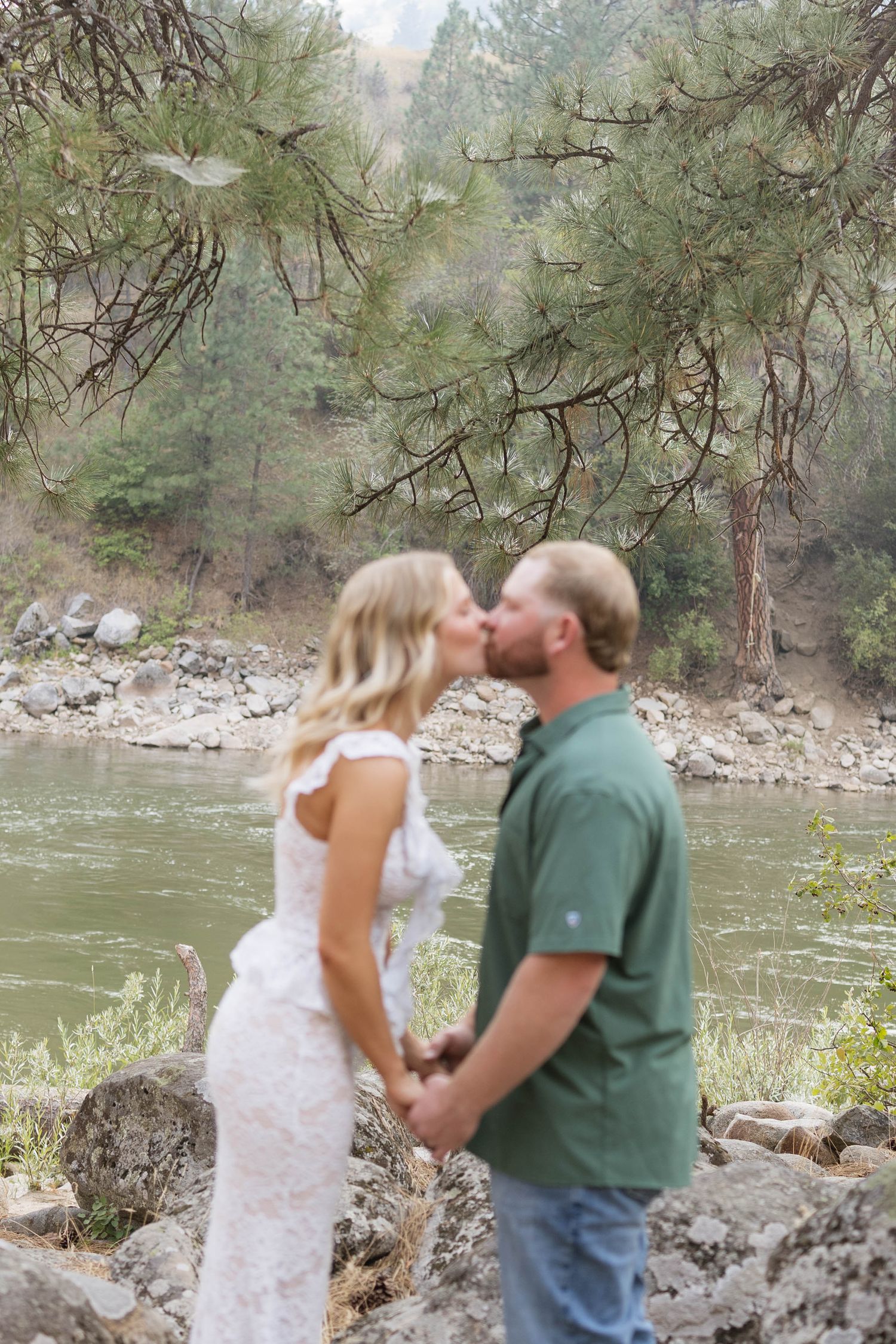 Couple in a white lace dress and green button-up shirt kiss while holding hands on a rocky riverbank surrounded by pine trees.