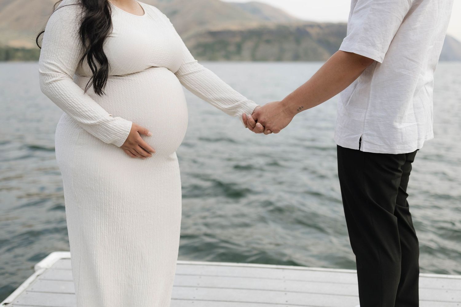 Pregnant woman in white dress holds partner's hand on dock with mountain lake backdrop.