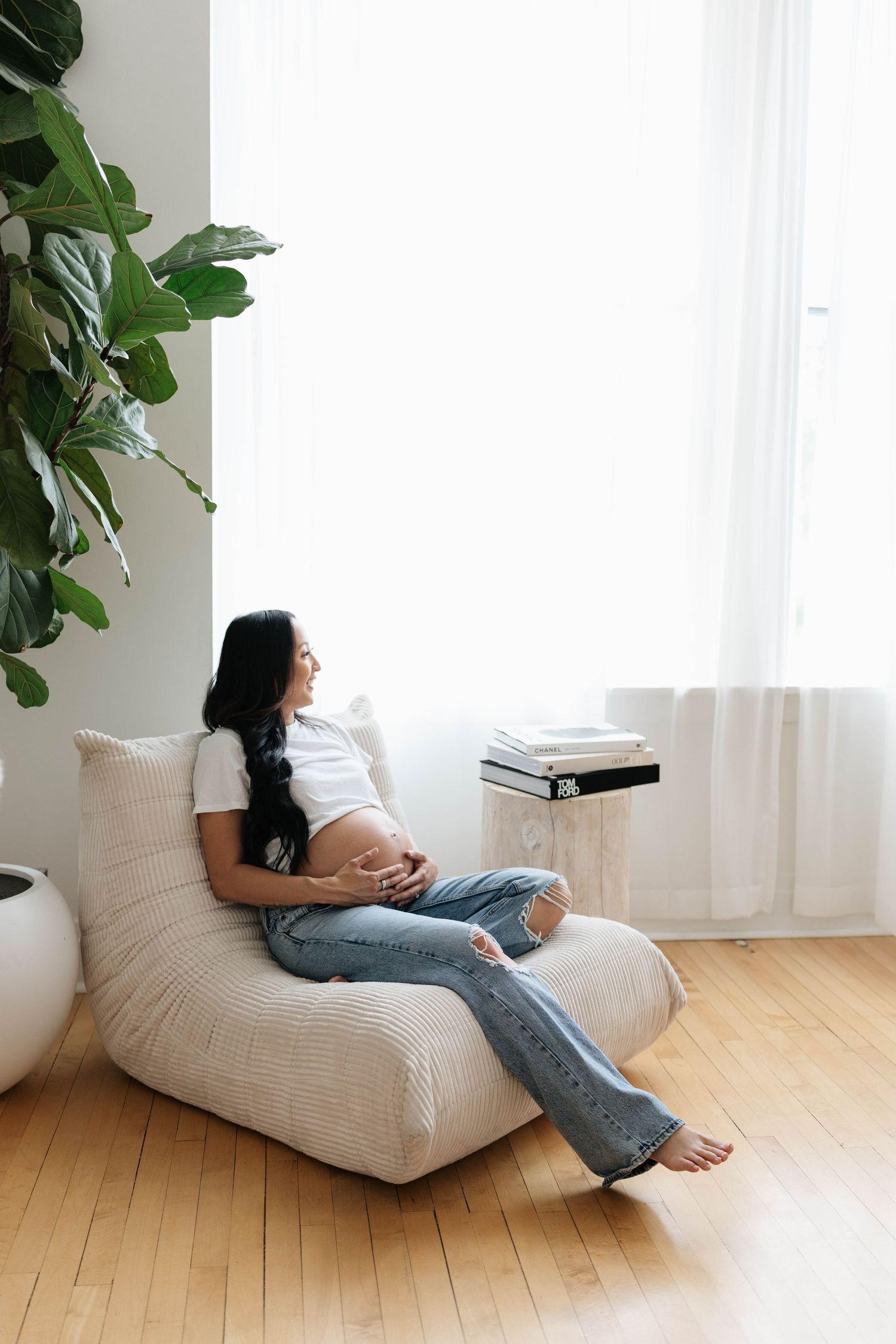 Pregnant woman in white tee and jeans sits in a cream lounge chair, cradling her belly near a bright window.