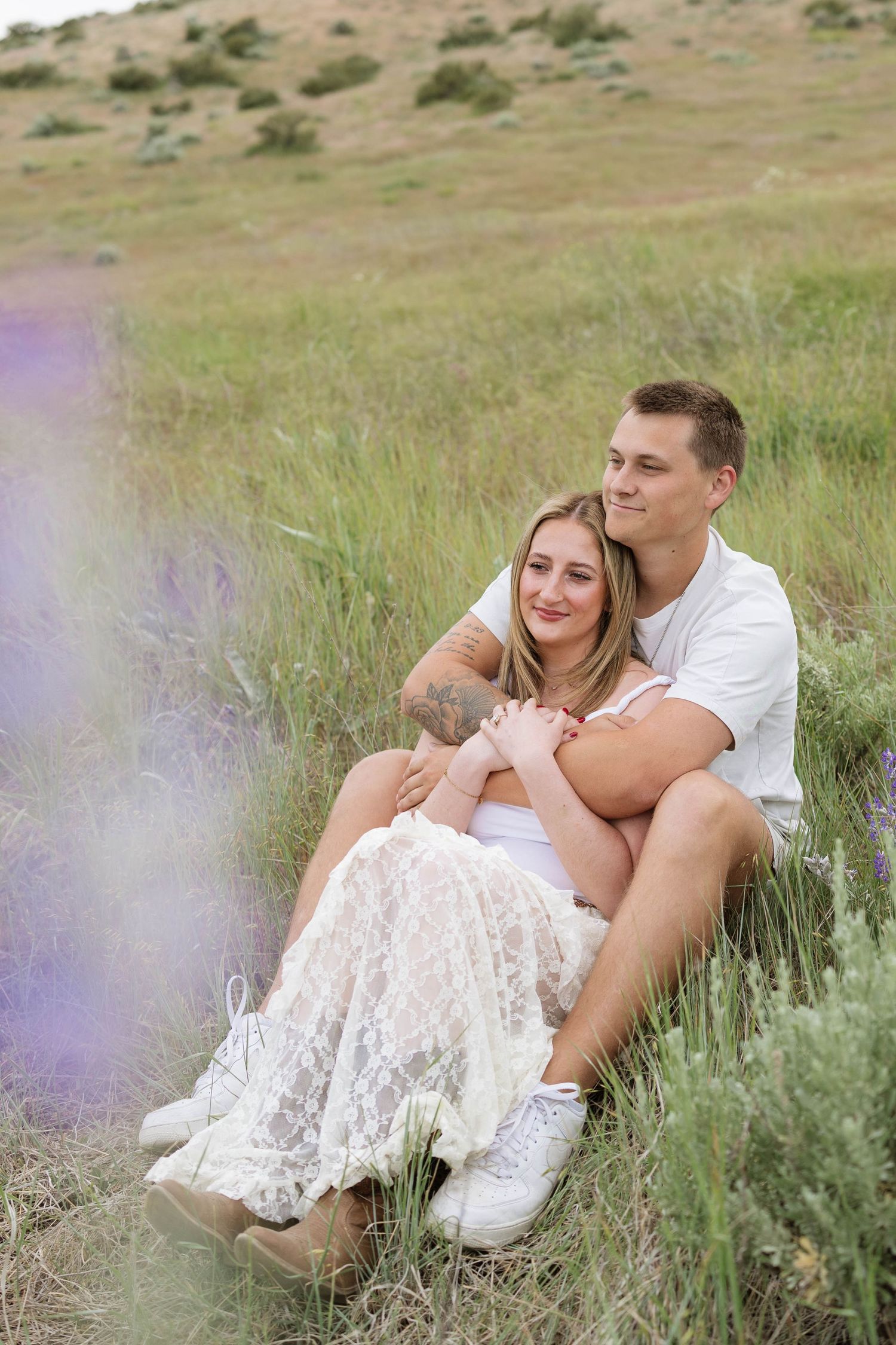 Couple sitting together in a grassy field, woman in white lace skirt leaning against man in white shirt.