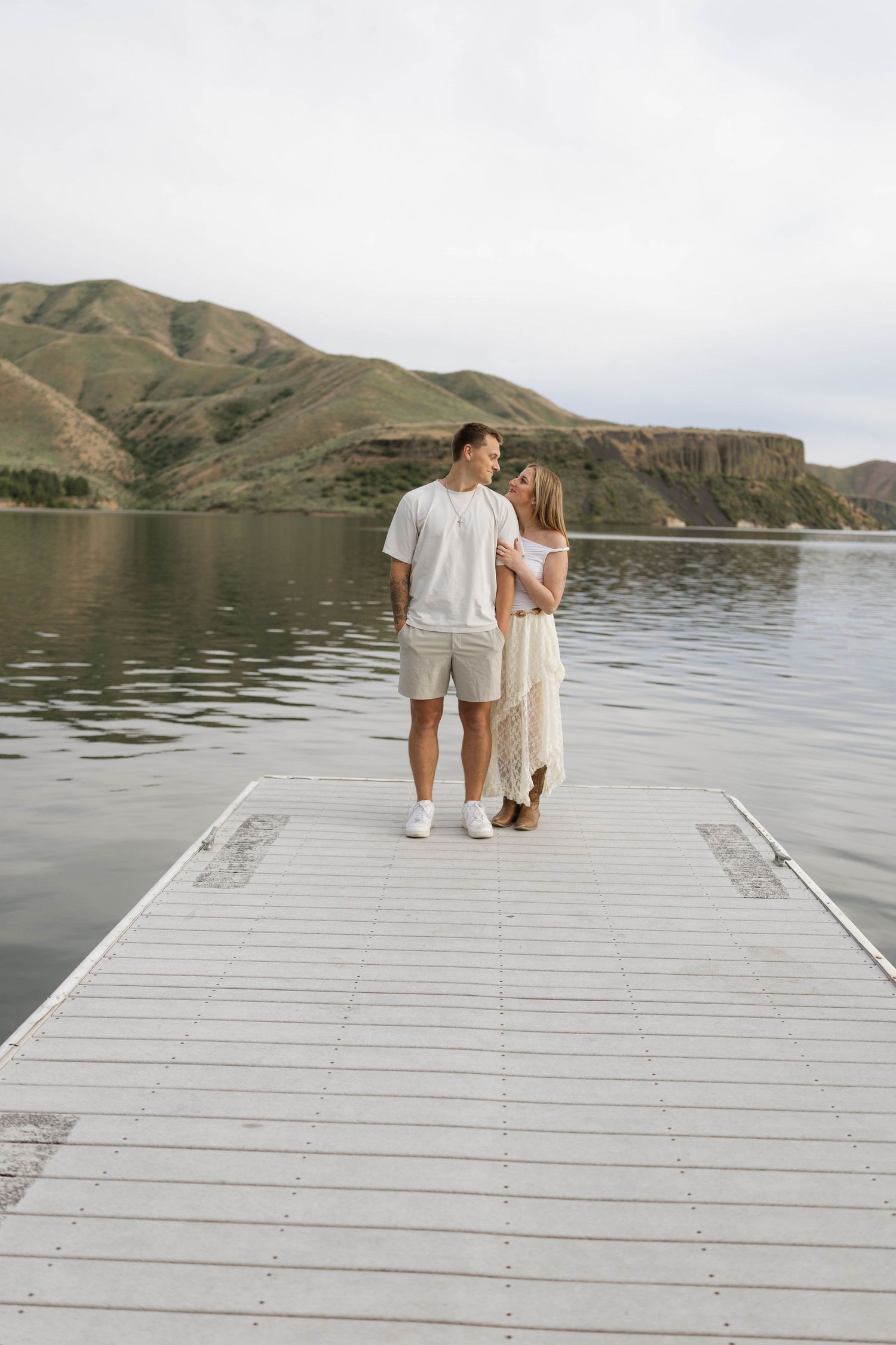Couple standing on a white dock by a calm lake with green hills in the background on a cloudy day.