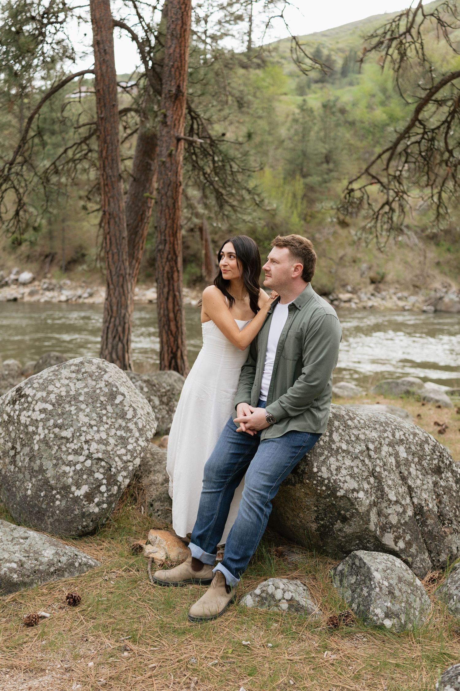 Couple poses on rocks by a river, woman in white dress embraces man in green jacket and jeans, surrounded by pine trees.