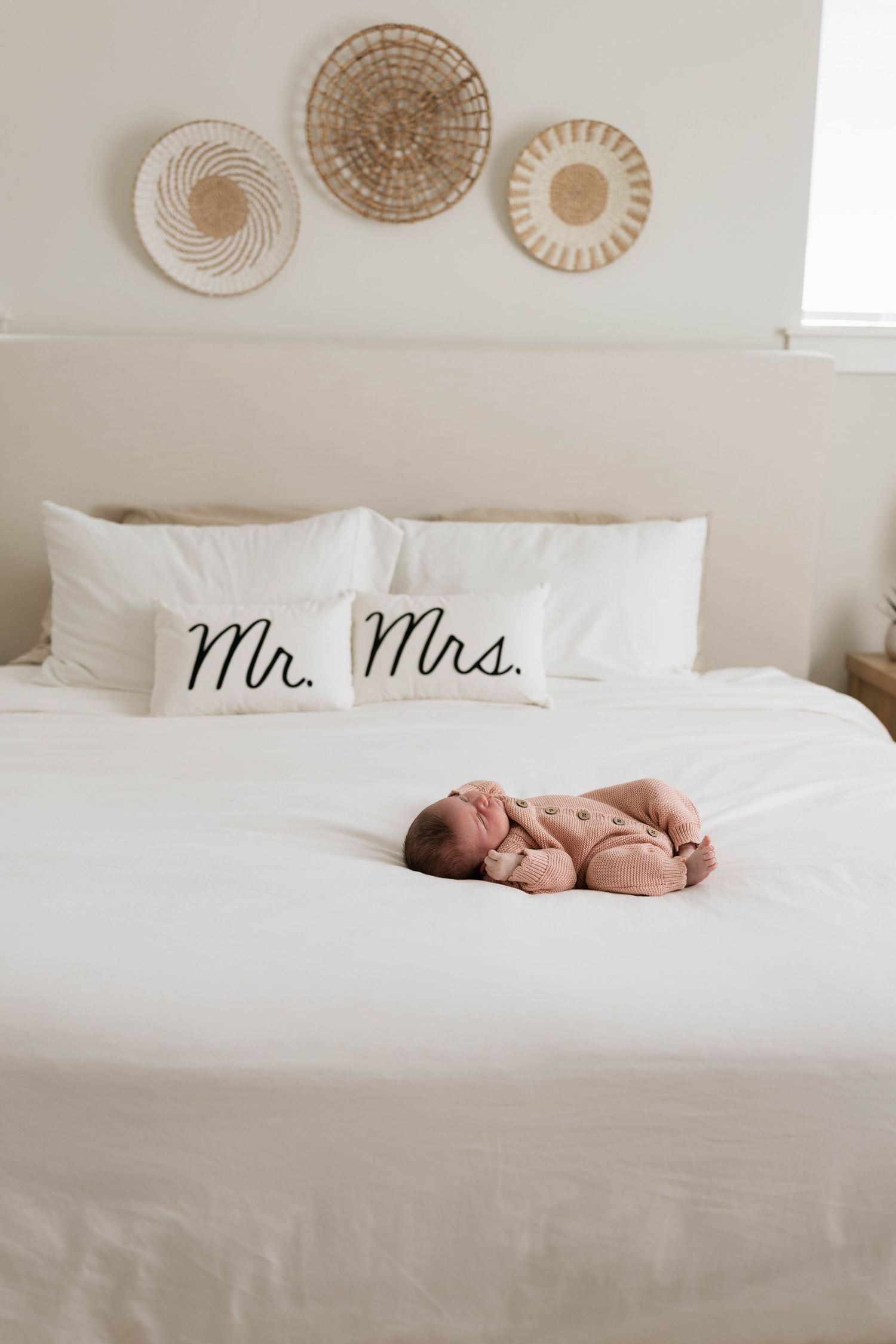 Newborn baby sleeping on white bed with Mr. and Mrs. pillows and woven wall decor in a bright bedroom.