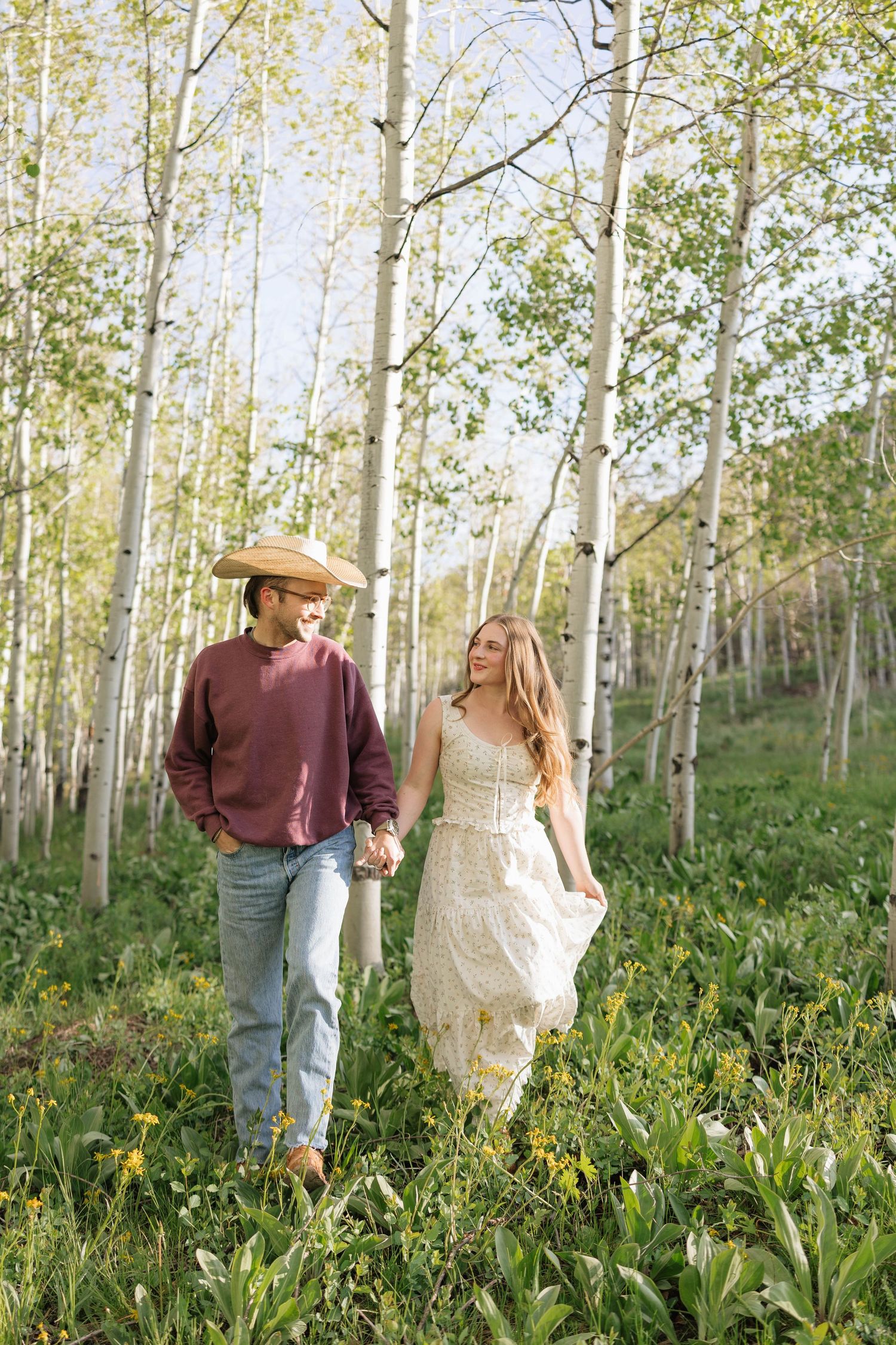 Couple holding hands walking through aspen grove in summer, woman in white lace dress, man in cowboy hat.