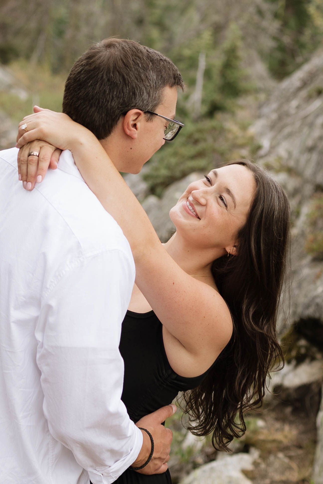 A smiling woman with long dark hair wraps her arms around a man in a white shirt during an outdoor couple session among rocks and trees.