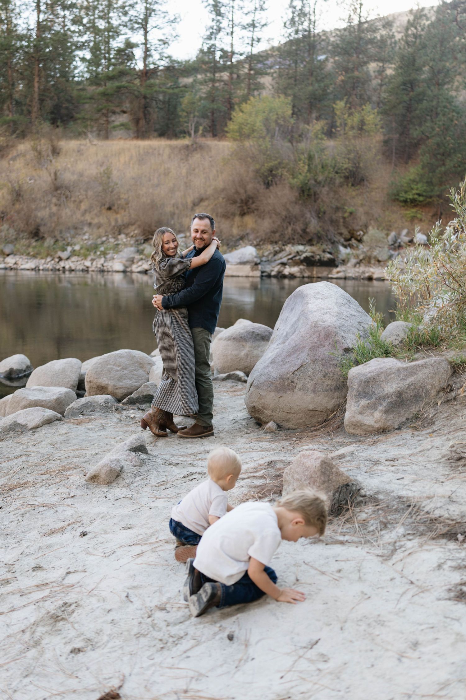 A couple embraces on a rocky riverbank while two young children in white shirts play in the sand during a family photo session.