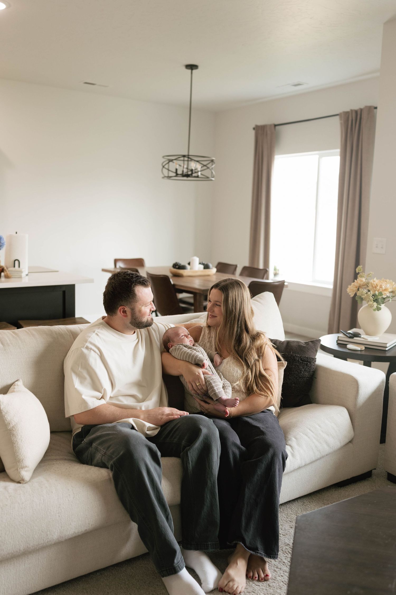 A couple sits on a white sofa, smiling at their newborn baby in a bright, modern living room.