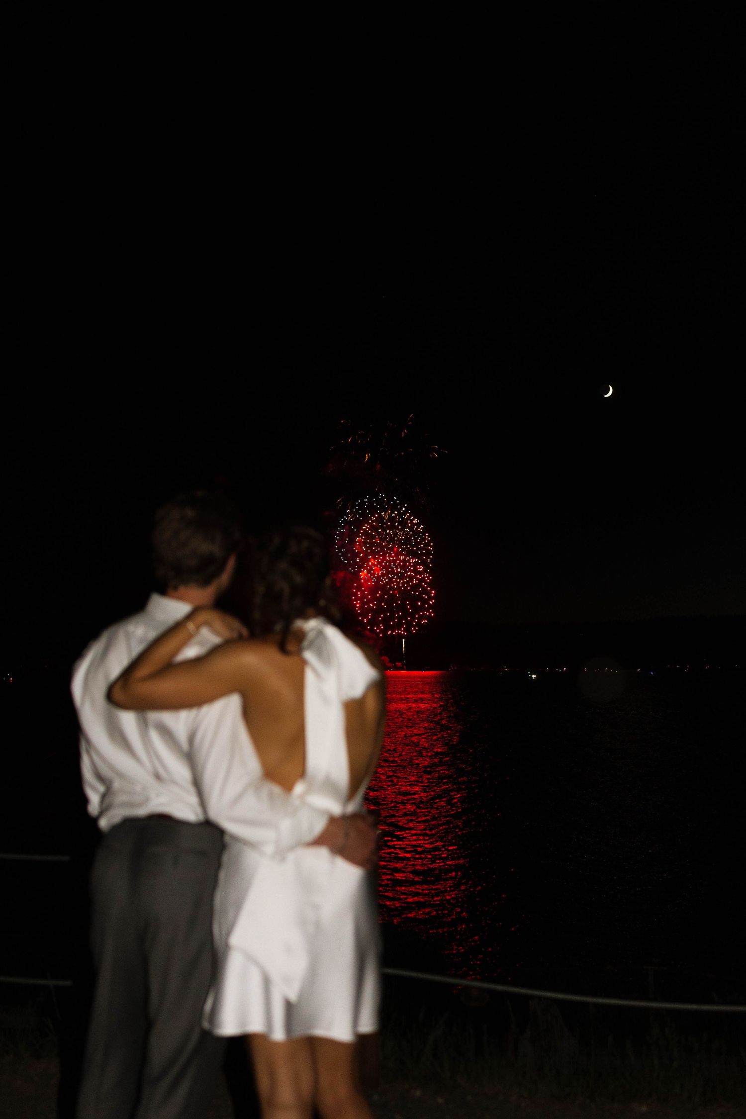 A couple in formal attire embraces while watching red fireworks illuminate the night sky in the distance.