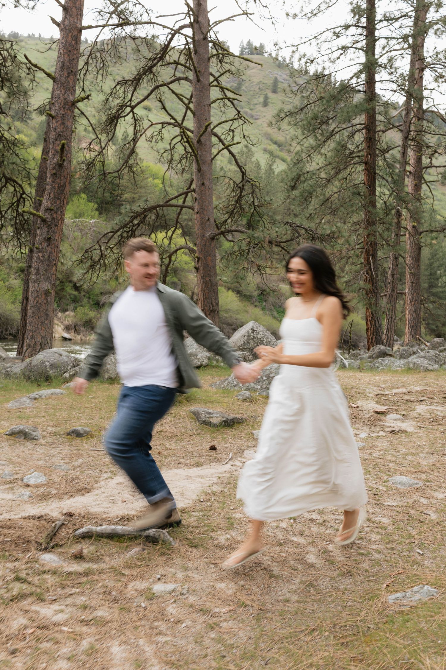 A joyful couple runs hand-in-hand through a forest clearing, the woman in a white dress and man in jeans and green jacket.
